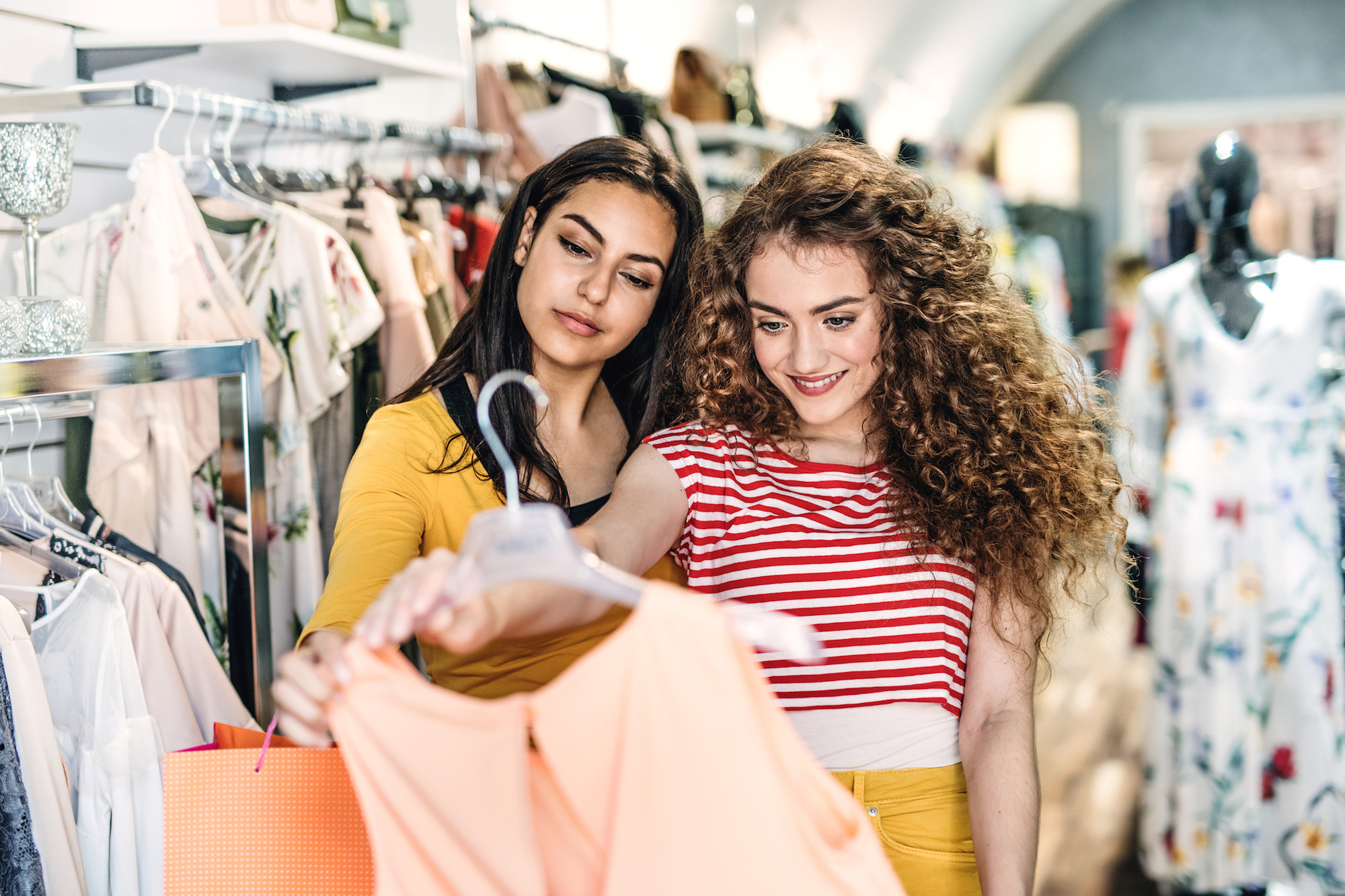 Two female teenager friends standing inside in the shop, holding and looking at dress.