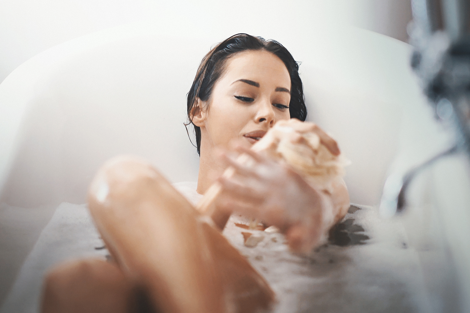 Closeup of attractive mid 20's woman taking a long bath in a bathtub. She's slowly scrubbing off with a bath sponge and not planning to get out some time soon.