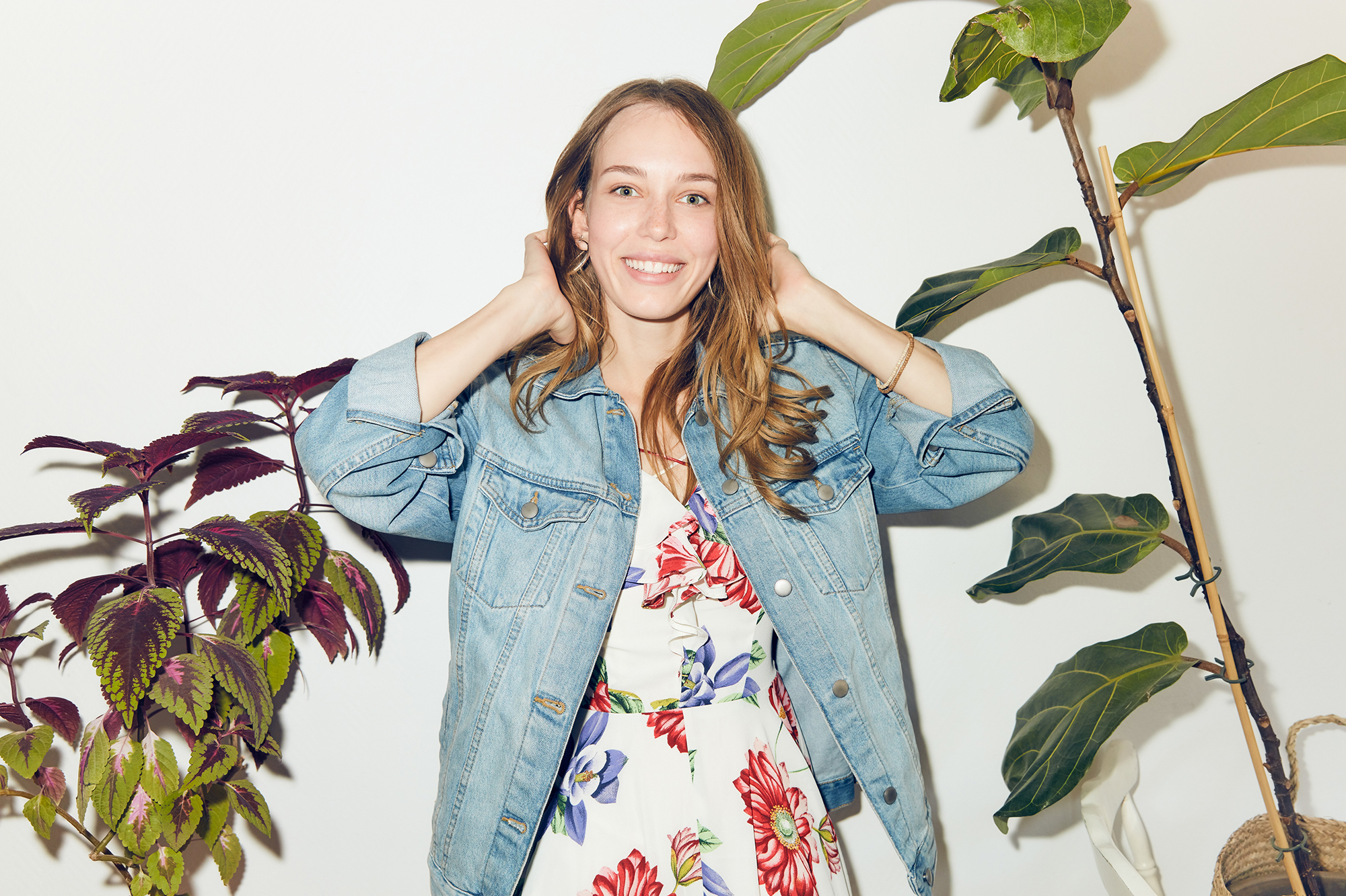Portrait of smiling young woman with hands in hair standing amidst plants against white wall at home