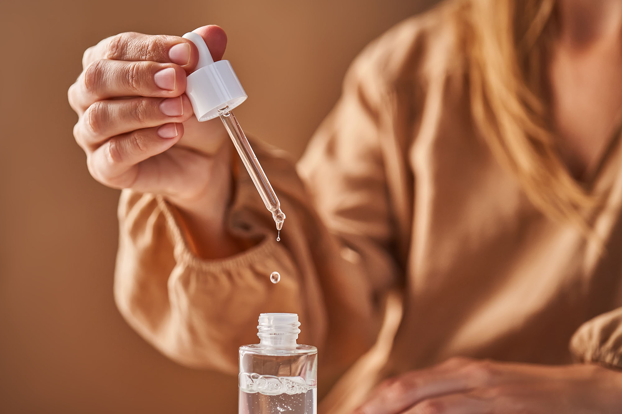 A girl in a beige dress holds a pipette with a drop of serum in her hands. Horizontal.