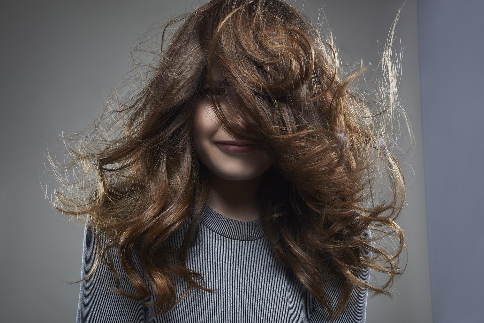 studio shot of a young female brunette in front of a gray background, wearing a gray outfit . her face half hidden behind her hair