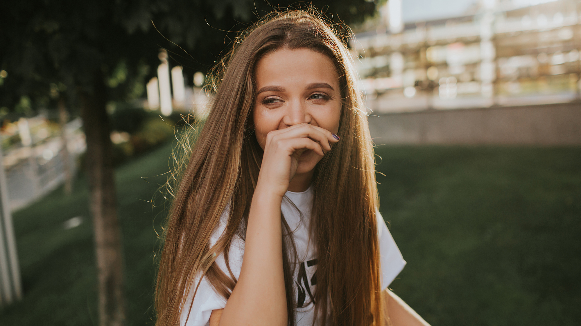 Stylish young blonde with extra long hair laughs cheerfully and looks away. She covers her mouth with palm. Sun illuminates her gently, well-groomed park is on the background.