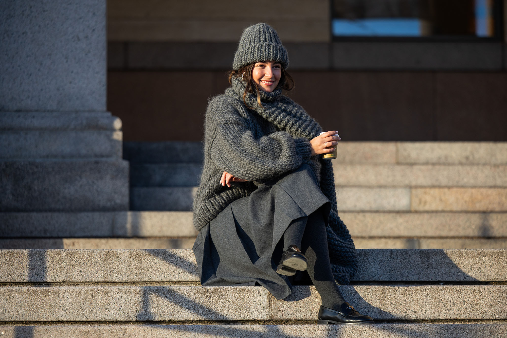 Inspiration: Benthe Liem sitting drinking coffee wears grey beanie, knit, scarf, skirt, black Prada bag outside Marimekko during the Copenhagen Fashion Week AW24 on February 01, 2024 in Copenhagen, Denmark. (Photo by Christian Vierig/Getty Images)