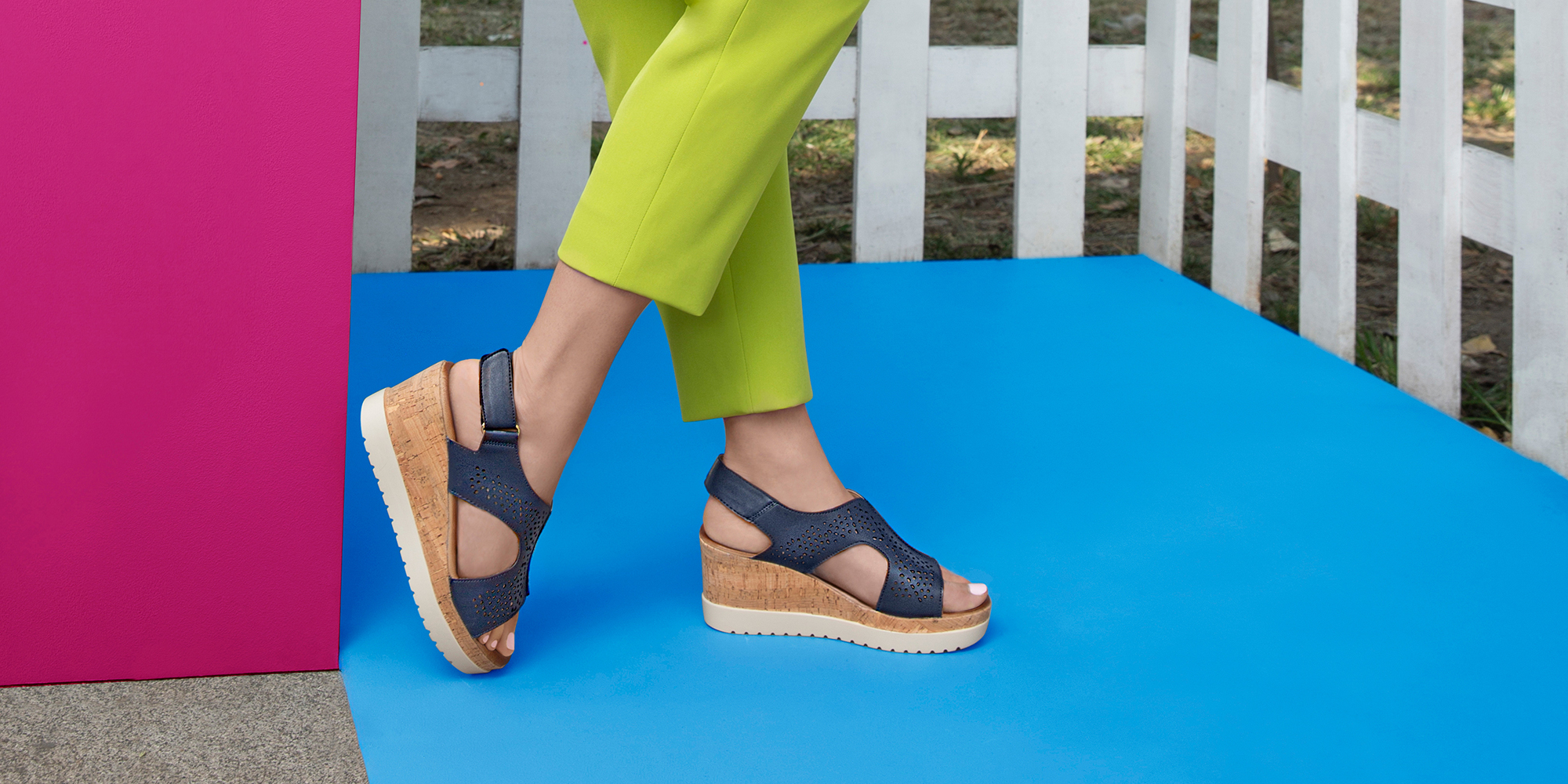 Outdoor scene featuring a woman modeling stylish navy blue wedge sandals, cork soles and perforated straps. stands on a bright blue floor, pink wall, wearing lime cropped pants, greenery background.
