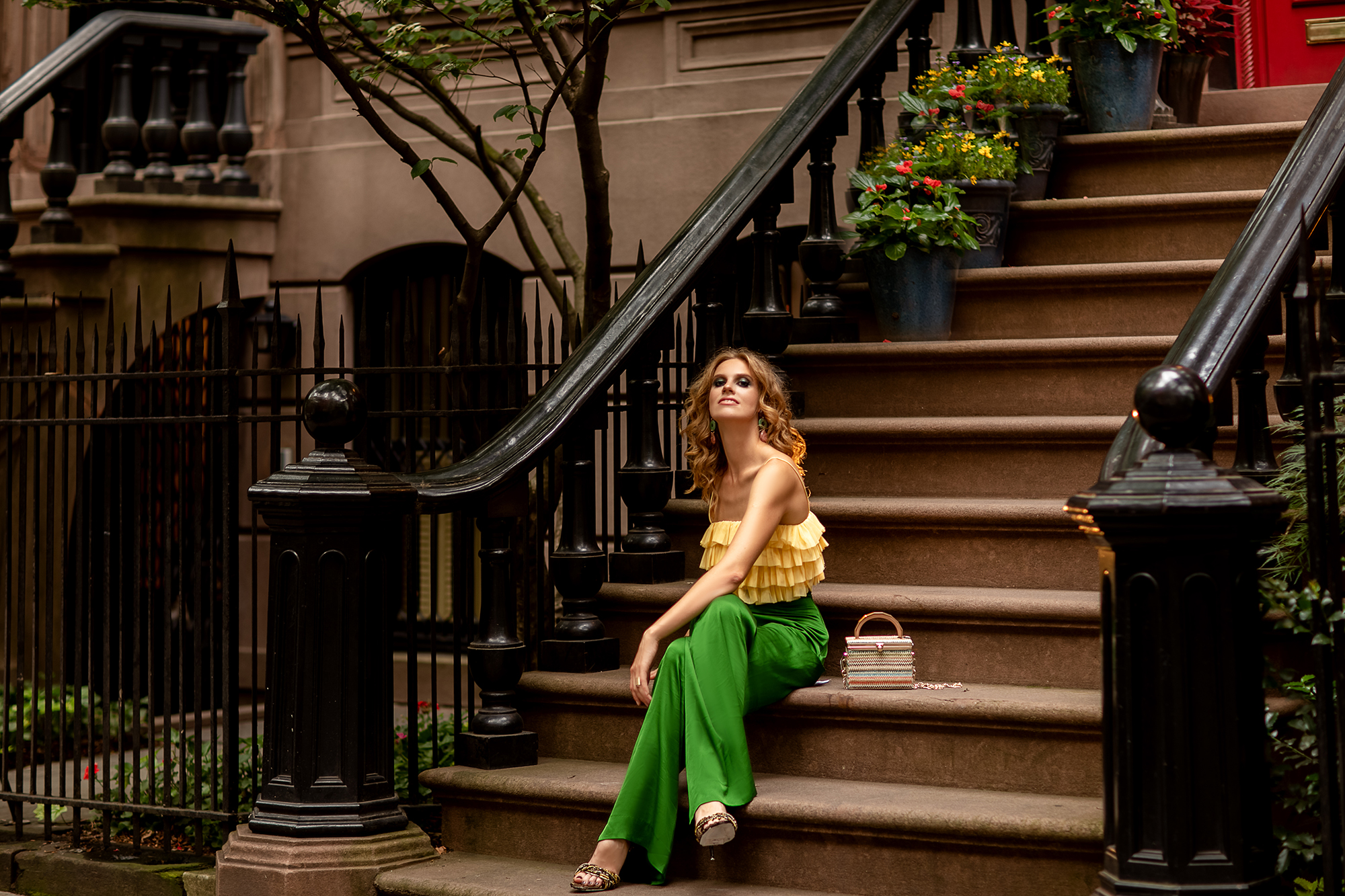 Red-haired young woman in stylish bright yellow top and green trousers elegant sandals sitting on the potted flowers porch stairs on Manhattan West village.