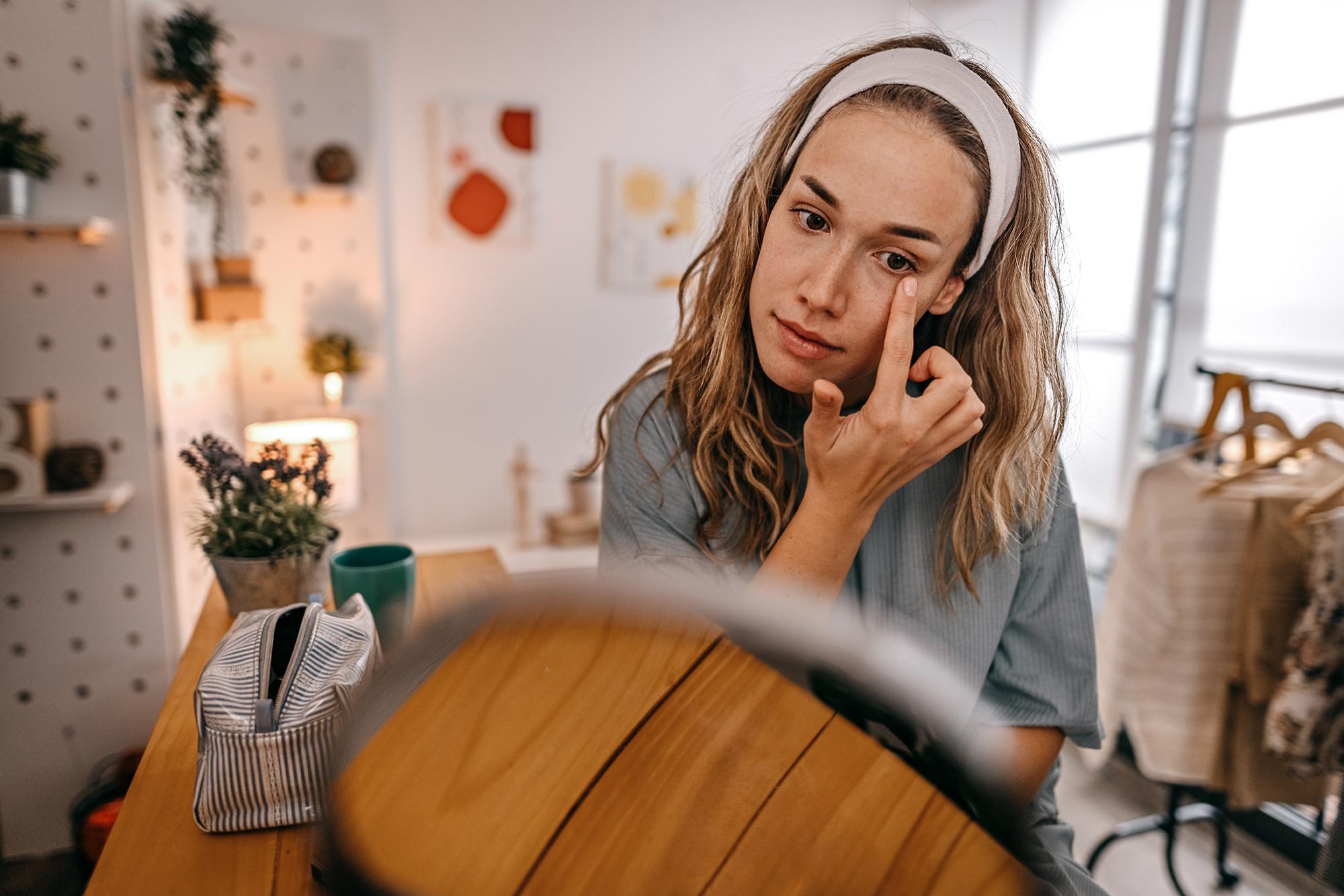 Young beautiful woman looking in mirror tightening her under eye skin with finger at home