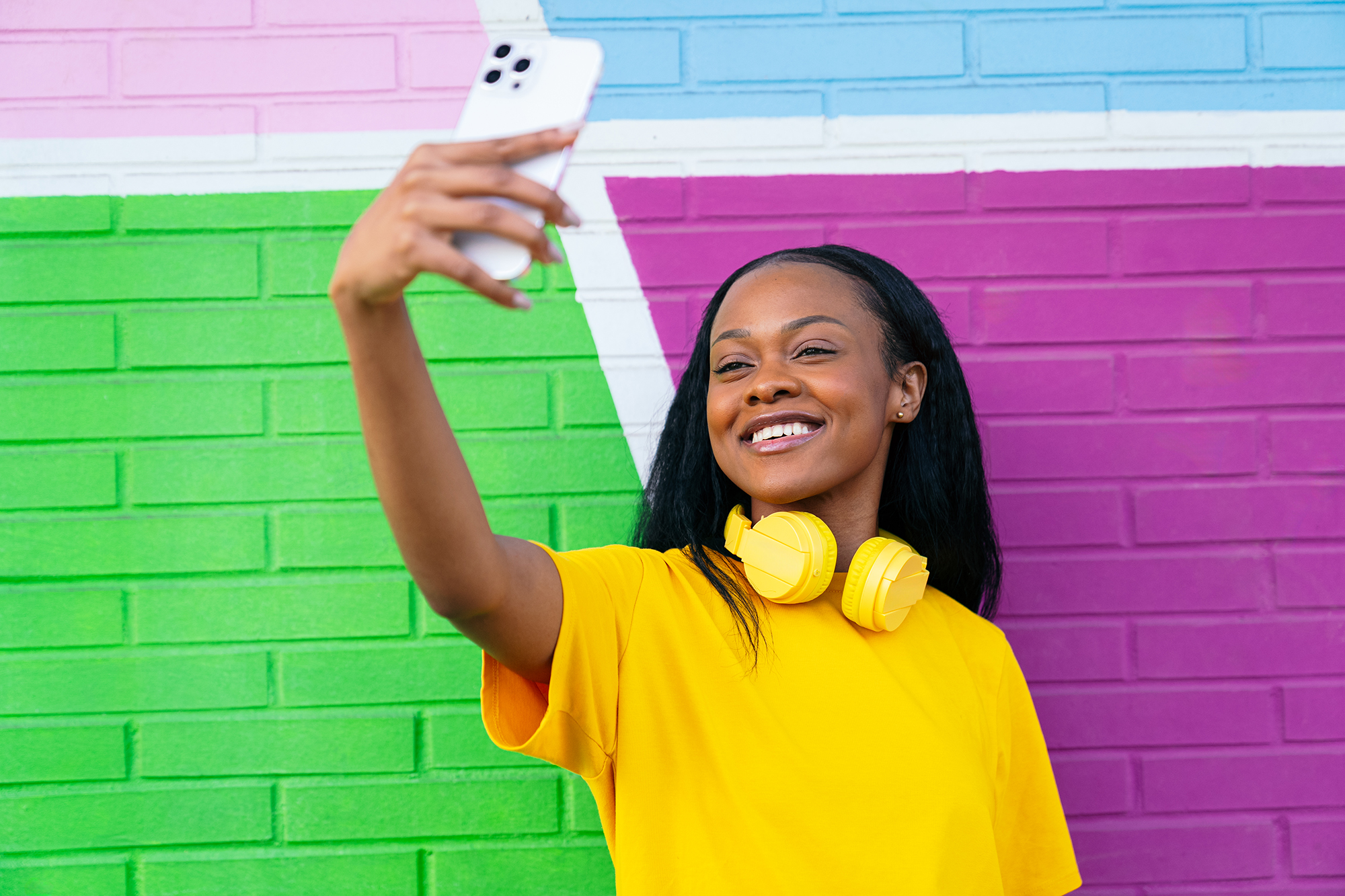 Brightly dressed woman with yellow headphones enjoys taking a selfie, with a vibrant urban wall behind.