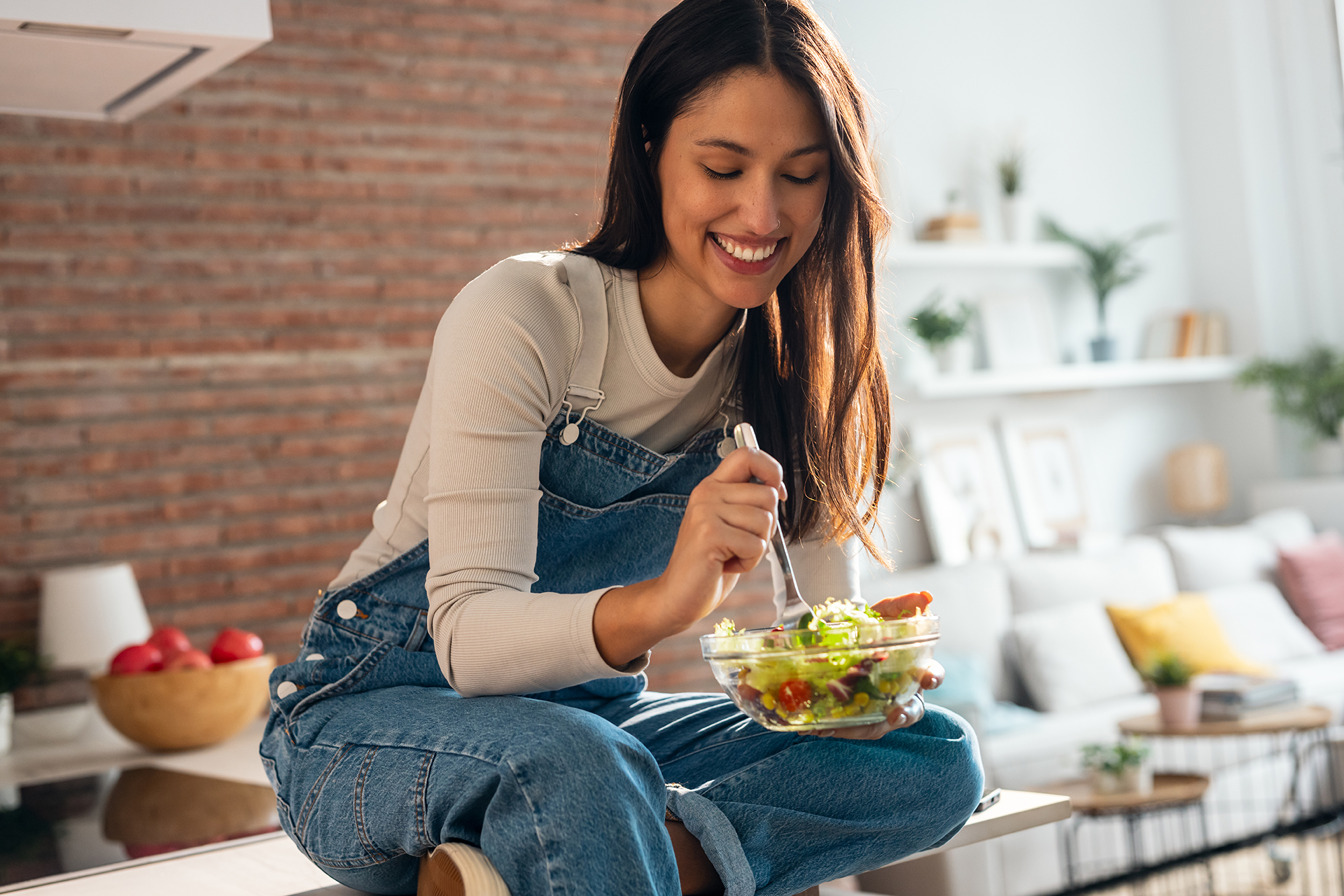 Shot of smiling woman eating healthy salad while sitting on the kitchen table at home.