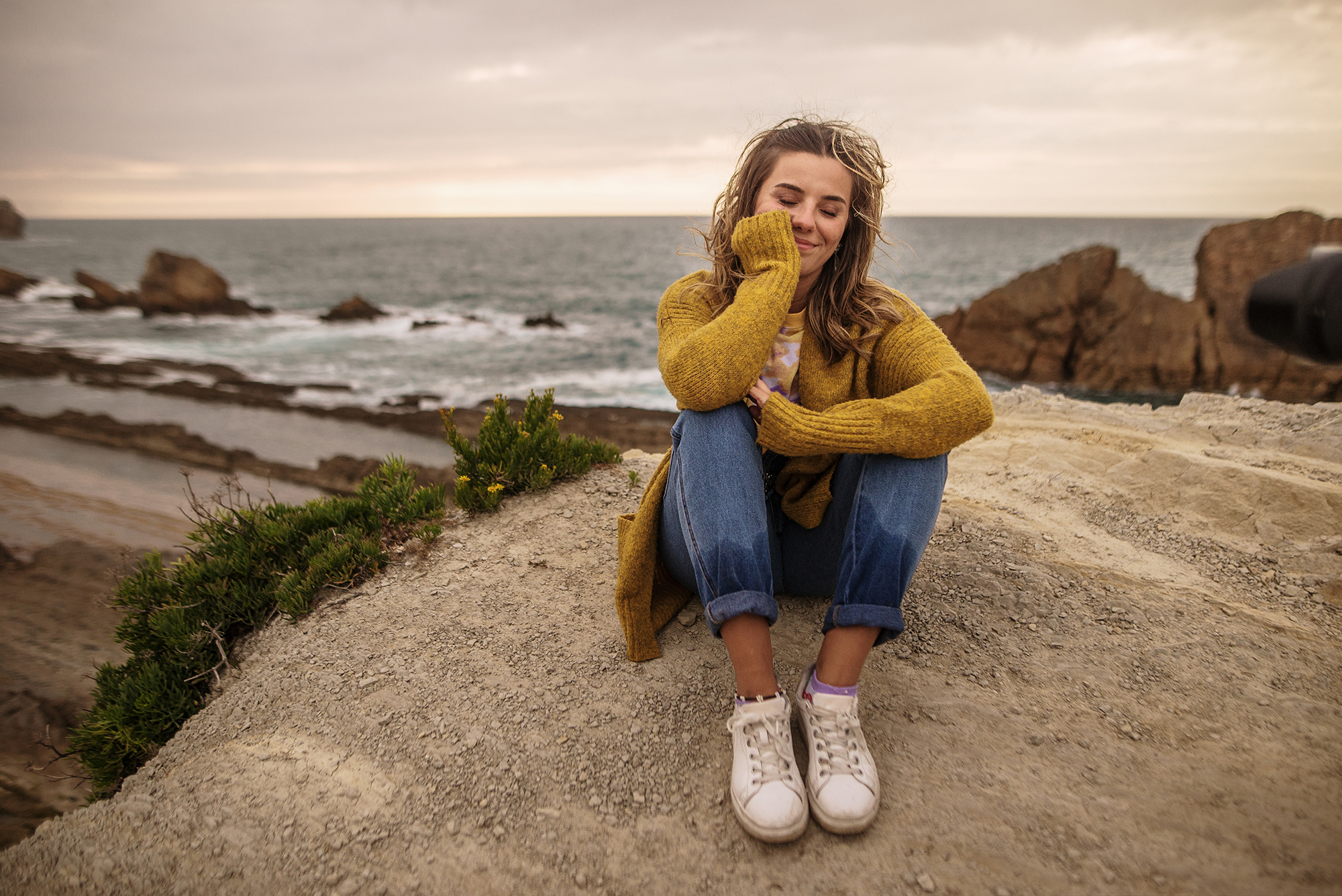 Young woman sitting and relaxing by the sea. She wears a yellow sweater and jeans