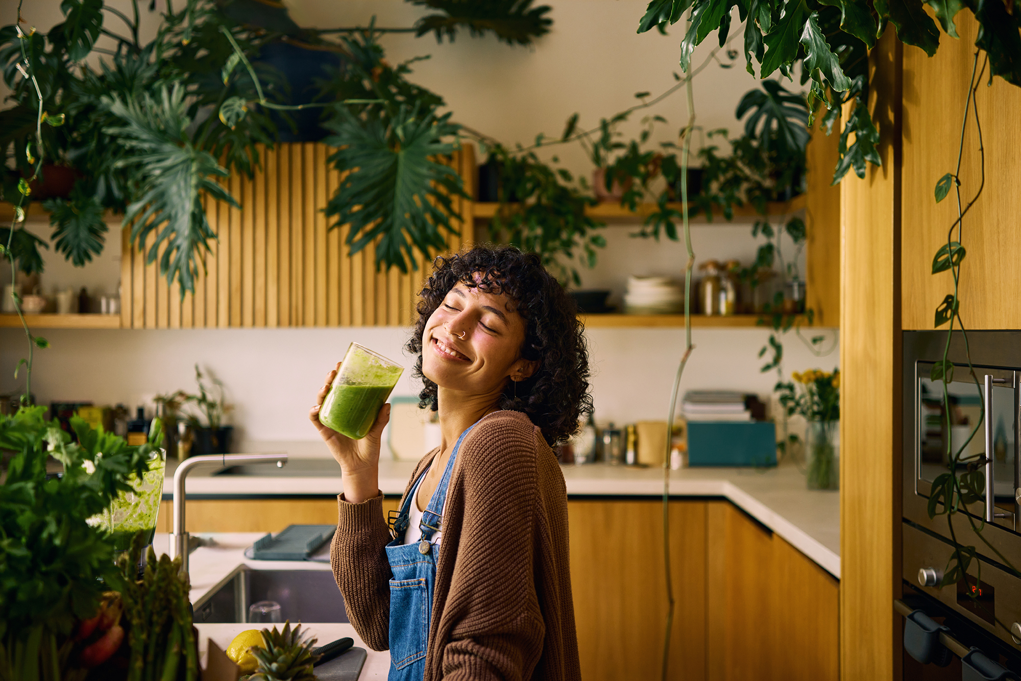Happy Woman Enjoying A Fresh Green Smoothie In A Sunny Kitchen Decorated With Indoor Plants