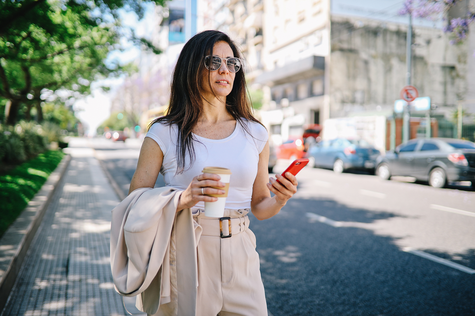 Fashionable woman walking on the street of Buenos Aires, looking for a taxi by using the mobile app on her smartphone.