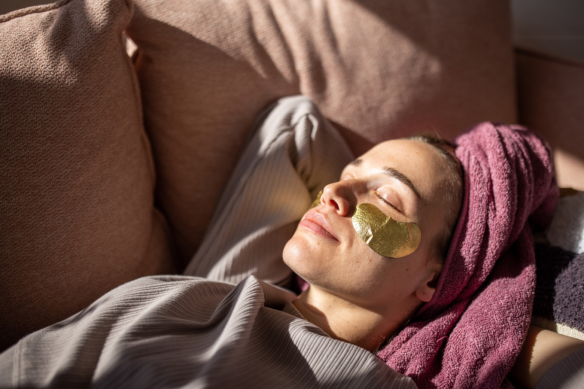 Young woman with eyes closed relaxing on sofa with towel wrapped around hair and gel patches under eyes.