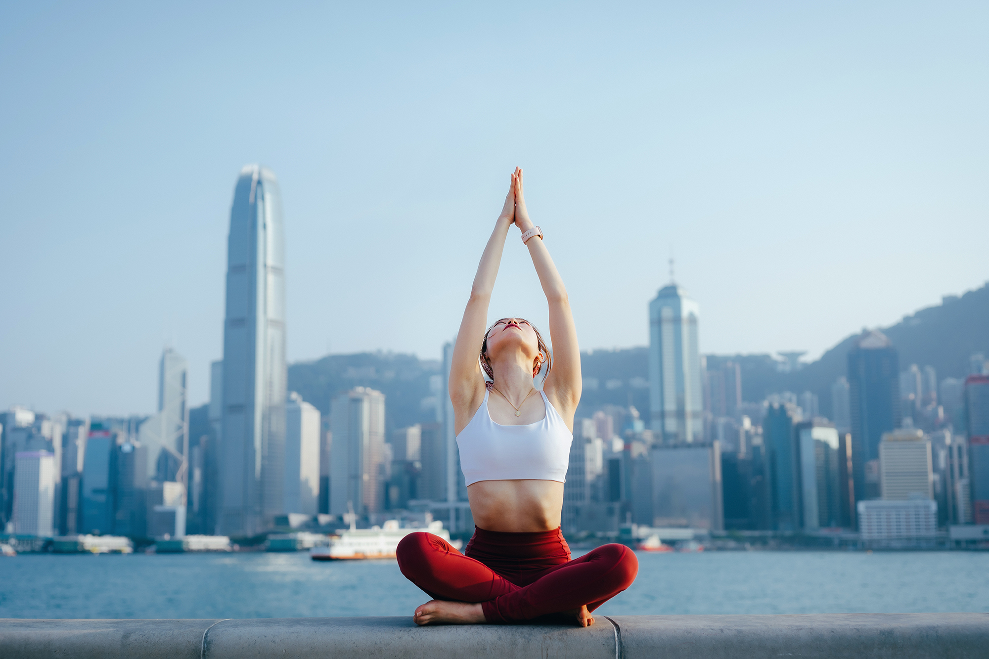 Young woman meditating outdoors, practicing mindfulness yoga in the morning, against spectacular Hong Kong city skyline by the promenade of Victoria harbour.