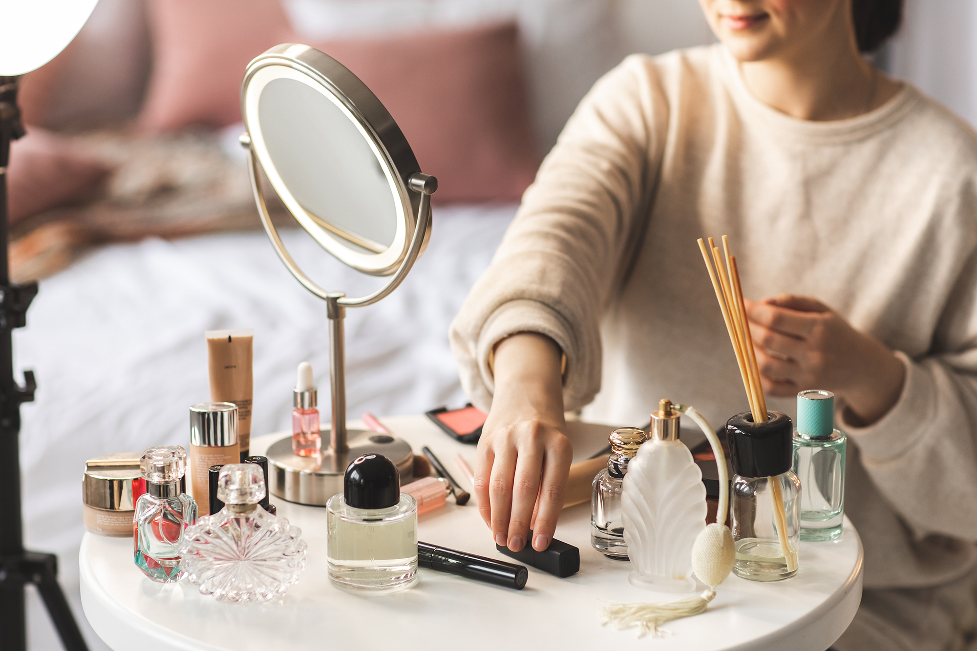 Cosmetics are laid out on a round white table. Beautiful little glass bottles and tubes with shiny lids, and some perfume. Woman is in the process of doing her make up.