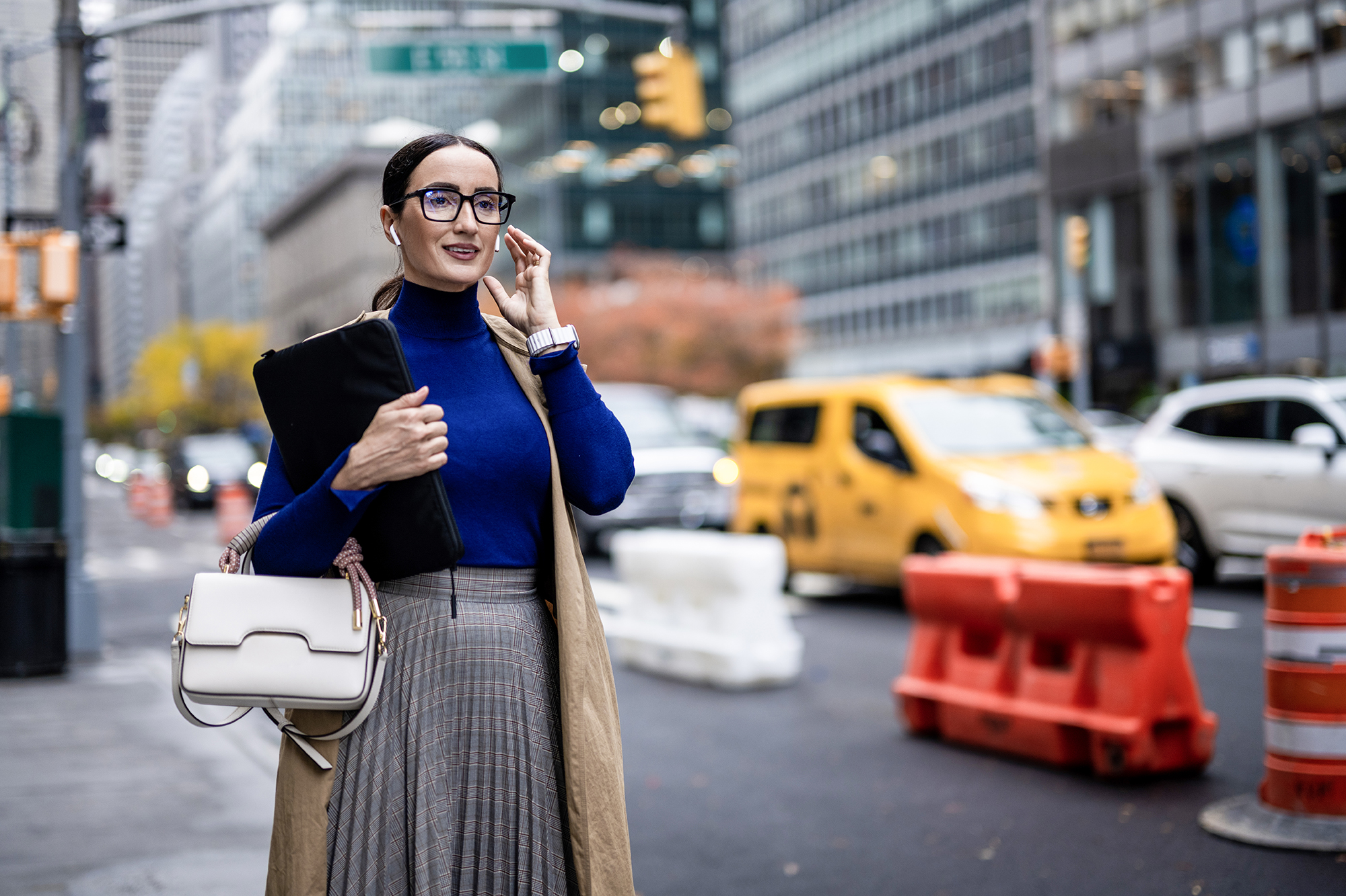 Smiling businesswoman with eyeglasses, wireless headphones and a laptop seen on the street of Manhattan using her wireless headphones after finishing work.