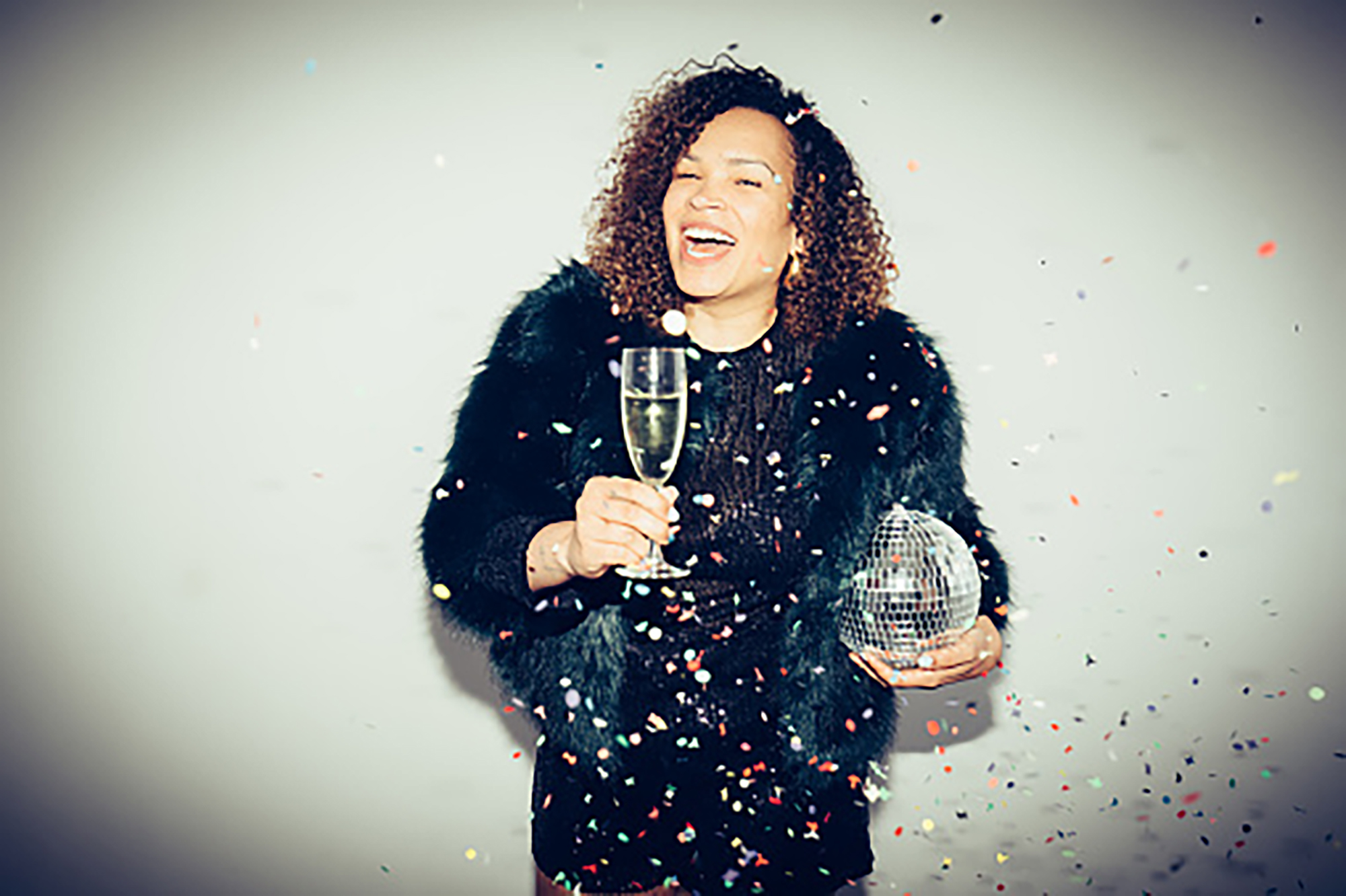 A smiling woman is holding a glass of champagne and a disco ball, wearing a black party dress amid falling confetti against a spot lit white background