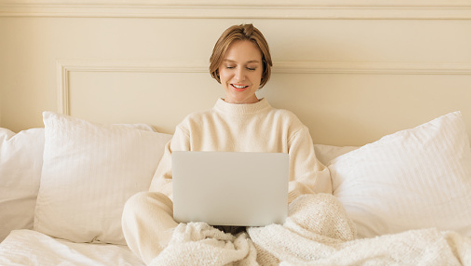 woman in bed on laptop in cream loungewear