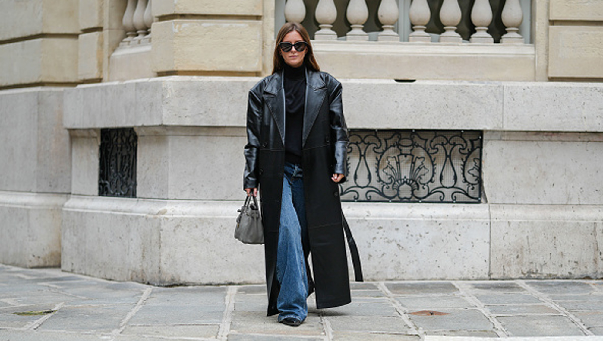 Diane Batoukina wears black Saint Laurent sunglasses, a black Tods turtleneck, a black faux leather long The Frankie Shop trench coat, a grey leather Birkin Hermes bag, blue denim jeans / pants, black leather Bershka shoes, during a street style fashion photo session, on October 18, 2024 in Paris, France.