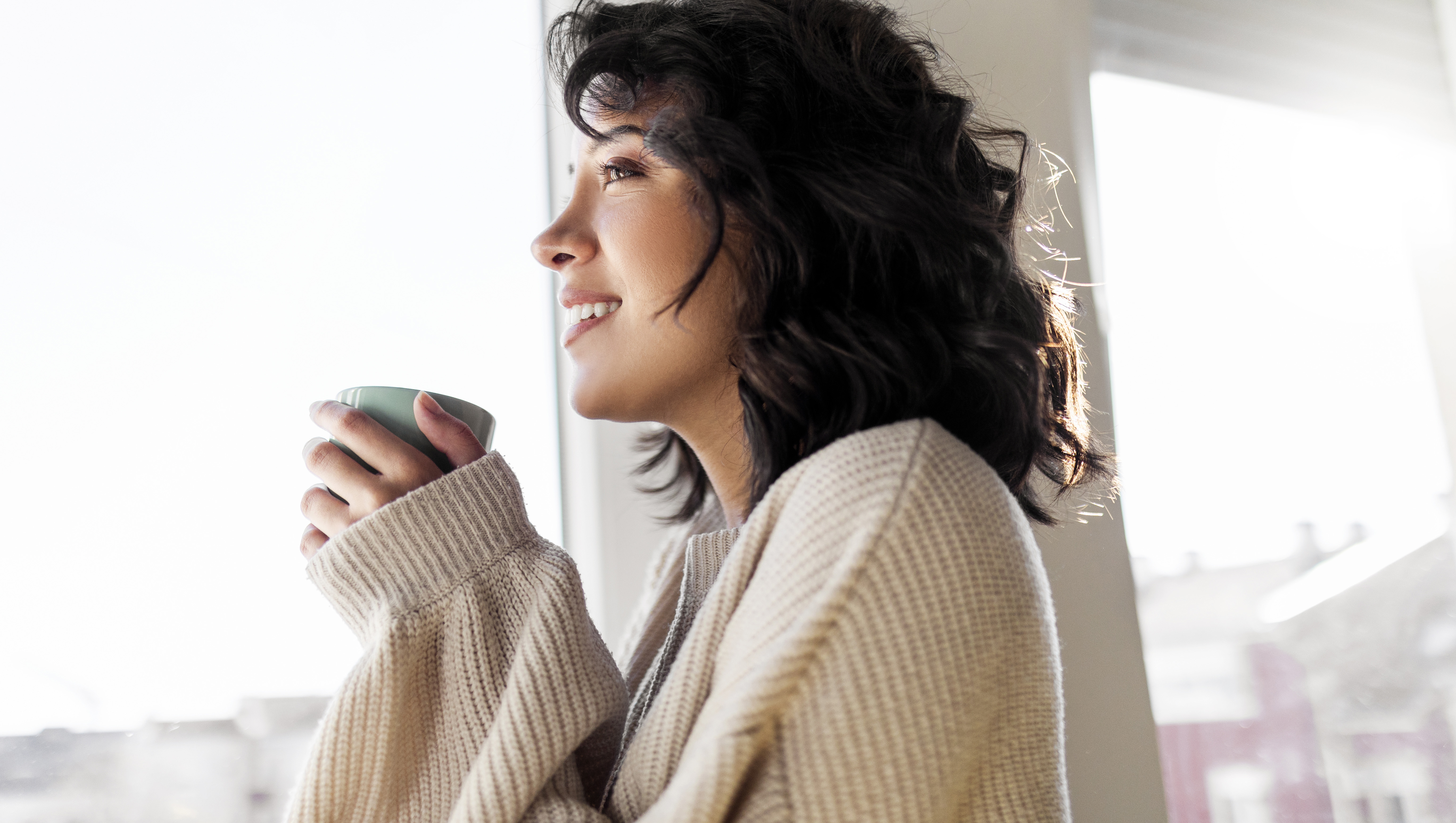 Beautiful young woman drinking cup of coffee while looking through window at home
