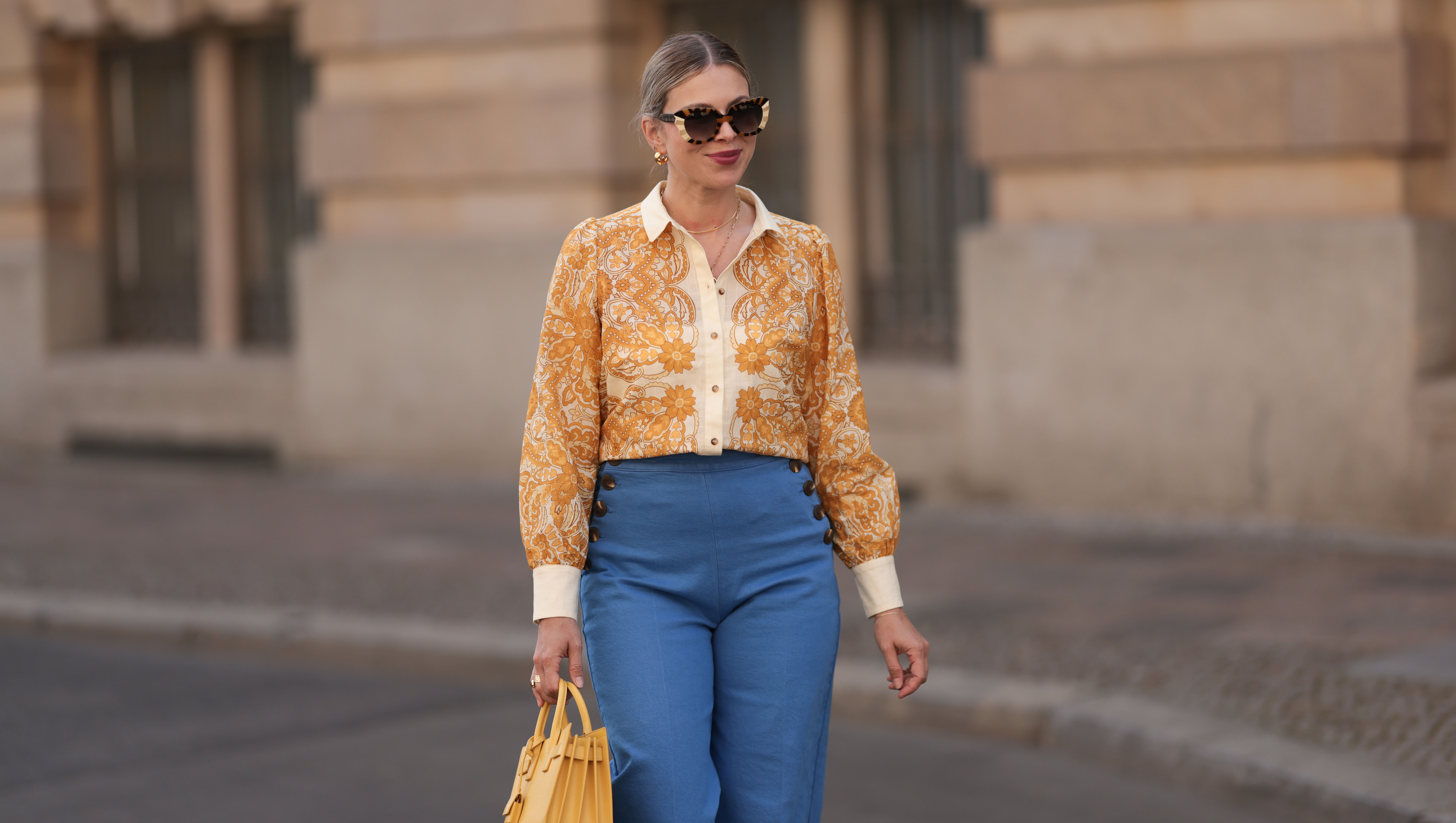 BERLIN, GERMANY - JUNE 07: Aline Kaplan is seen wearing large sunglasses with beige, brown and black pattern from Tibi; chunky golden hoop earrings; several golden necklaces; a creme-white blouse with floral orange pattern from Sézane; a blue high-waisted pair of trousers with two button borders on the hips from Sézane; golden rings and a yellow leather handbag from Saint Laurent on June 07, 2024 in Berlin, Germany.