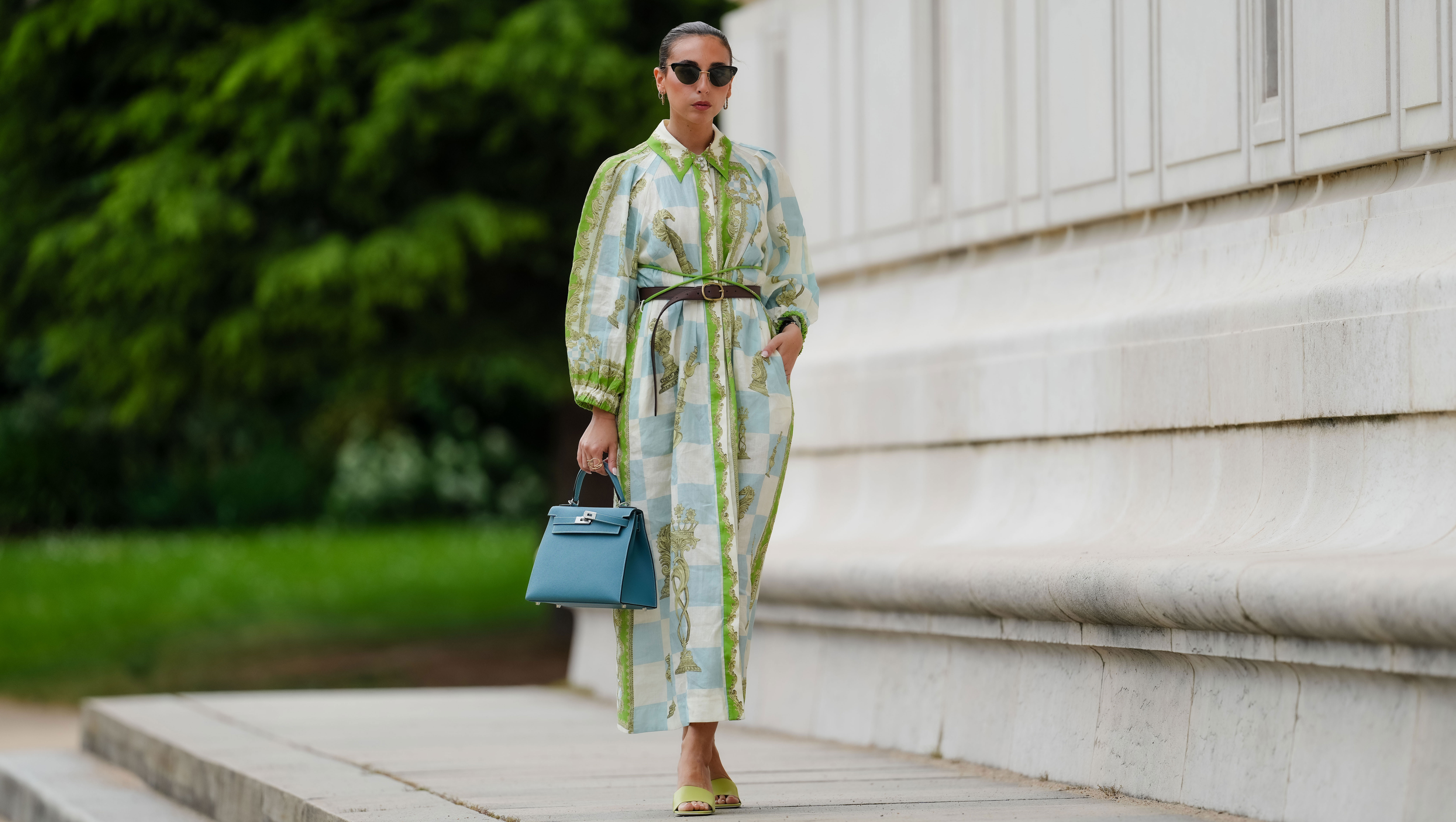 PARIS, FRANCE - MAY 12: Gabriella Berdugo wears sunglasses, earrings, a white blue and green checked long dress with long sleeves and floral print, a brown leather belt, a blue Kelly Hermes bag, pale pastel yellow mules / shoes with heart-shaped heels from Kat Maconie, during a street style fashion photo session, on May 12, 2024 in Paris, France.