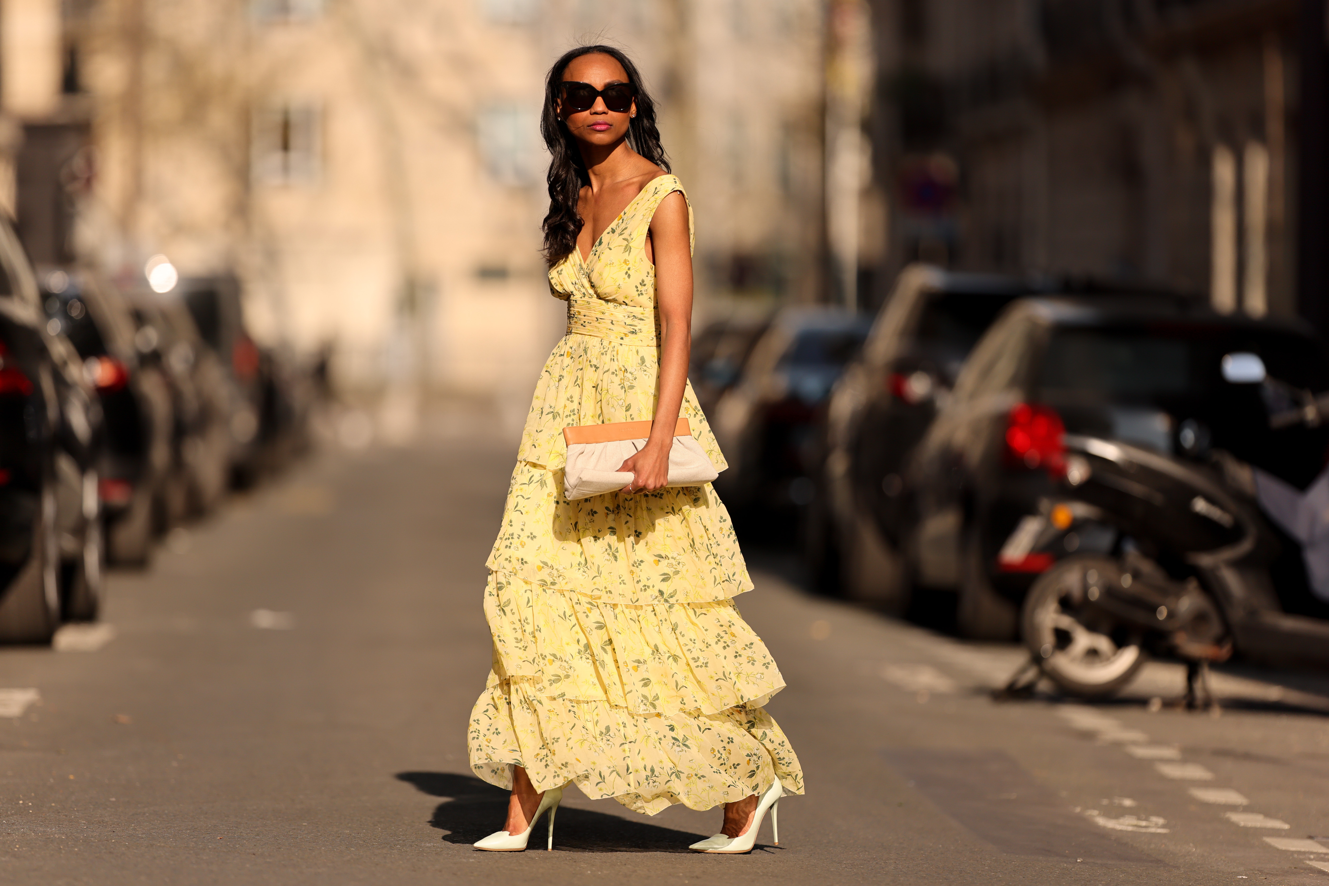 Emilie Joseph @in_fashionwetrust wears black sunglasses, a pale yellow with green and yellow flower print pattern V-neck / tank-top / backless / belted / ruffled maxi long dress, a beige fabric ruffled clutch, silver rings, pale yellow matte leather pointed pumps heels shoes, during a street style fashion photo session, on March 26, 2022 in Paris, France.