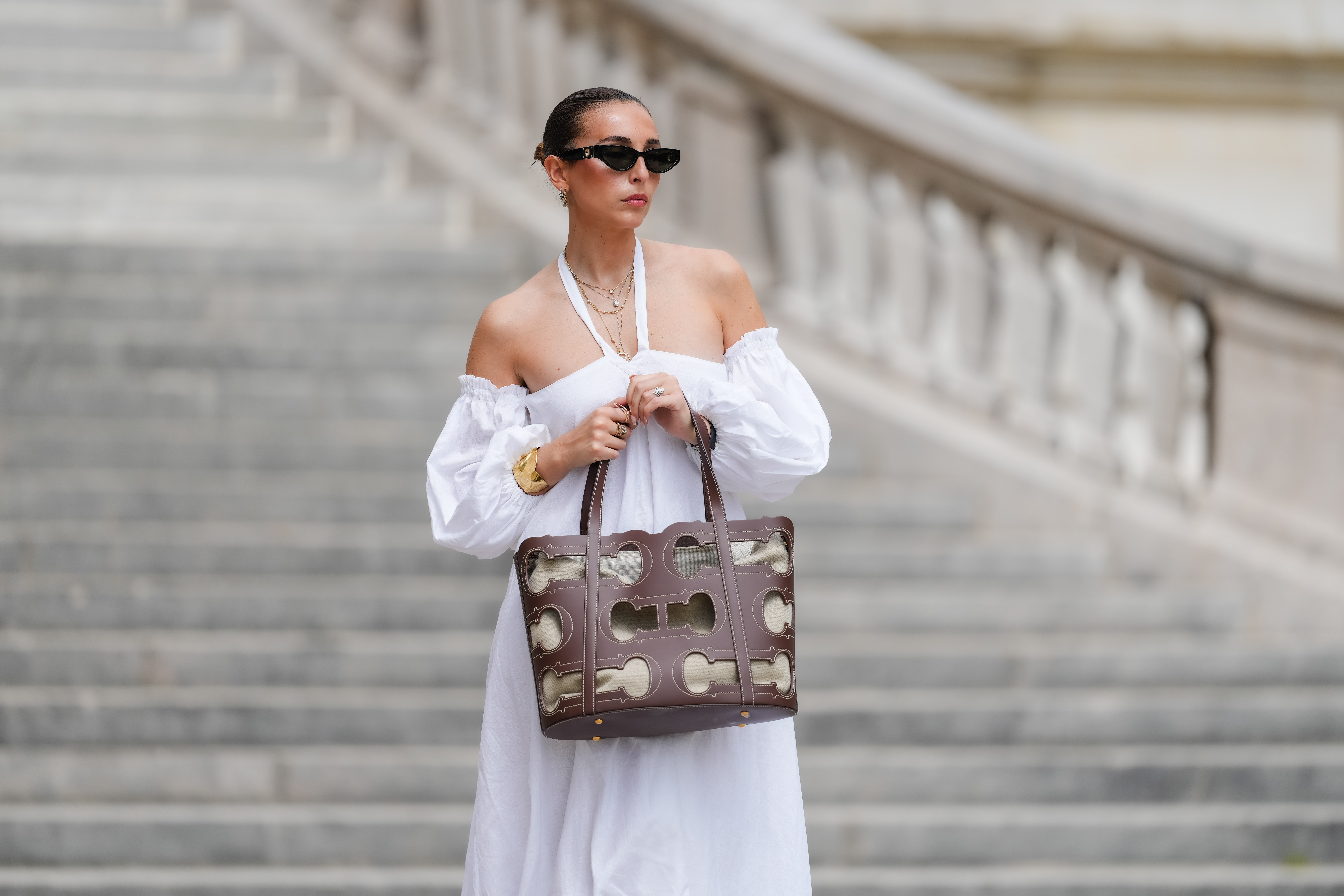 Gabriella Berdugo wears sunglasses, earrings, a white off-shoulder linen gathered dress from kalita with balloons / puff sleeves, a brown summer bag from Carolina Herrera in brown leather, during a street style fashion photo session, on May 12, 2024 in Paris, France.