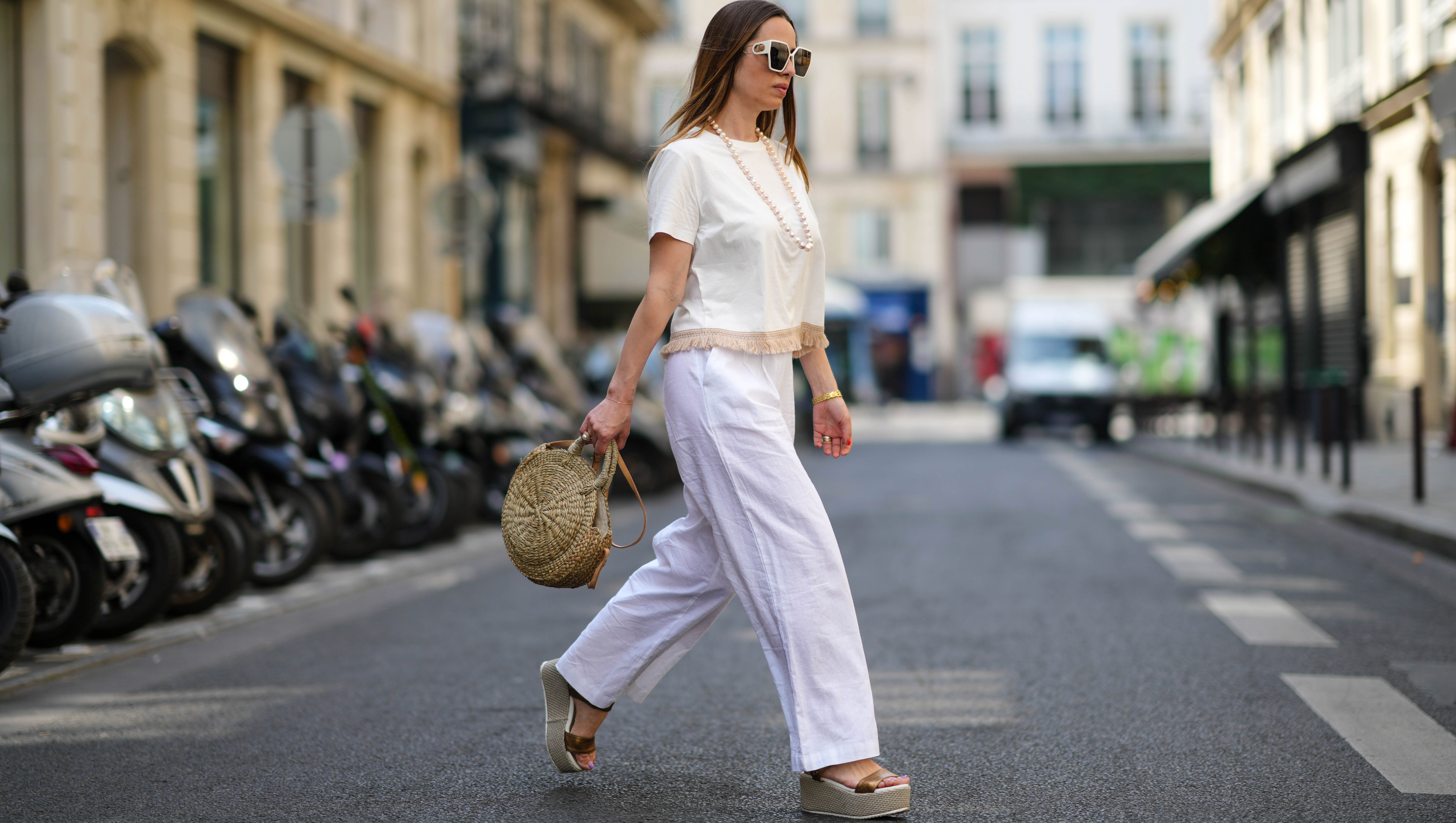 PARIS, FRANCE - MAY 10: Maria Rosaria Rizzo wears white sunglasses, a white pearls long necklace, a white latte t-shirt with fringed borders, a beige wickers handbag from Guerlain, a gold large bracelet, gold rings, high waist white wide legs linen pants, gold shiny leather strappy with wicker wedge heels sandals, during a street style fashion photo session, on May 10, 2022 in Paris, France.