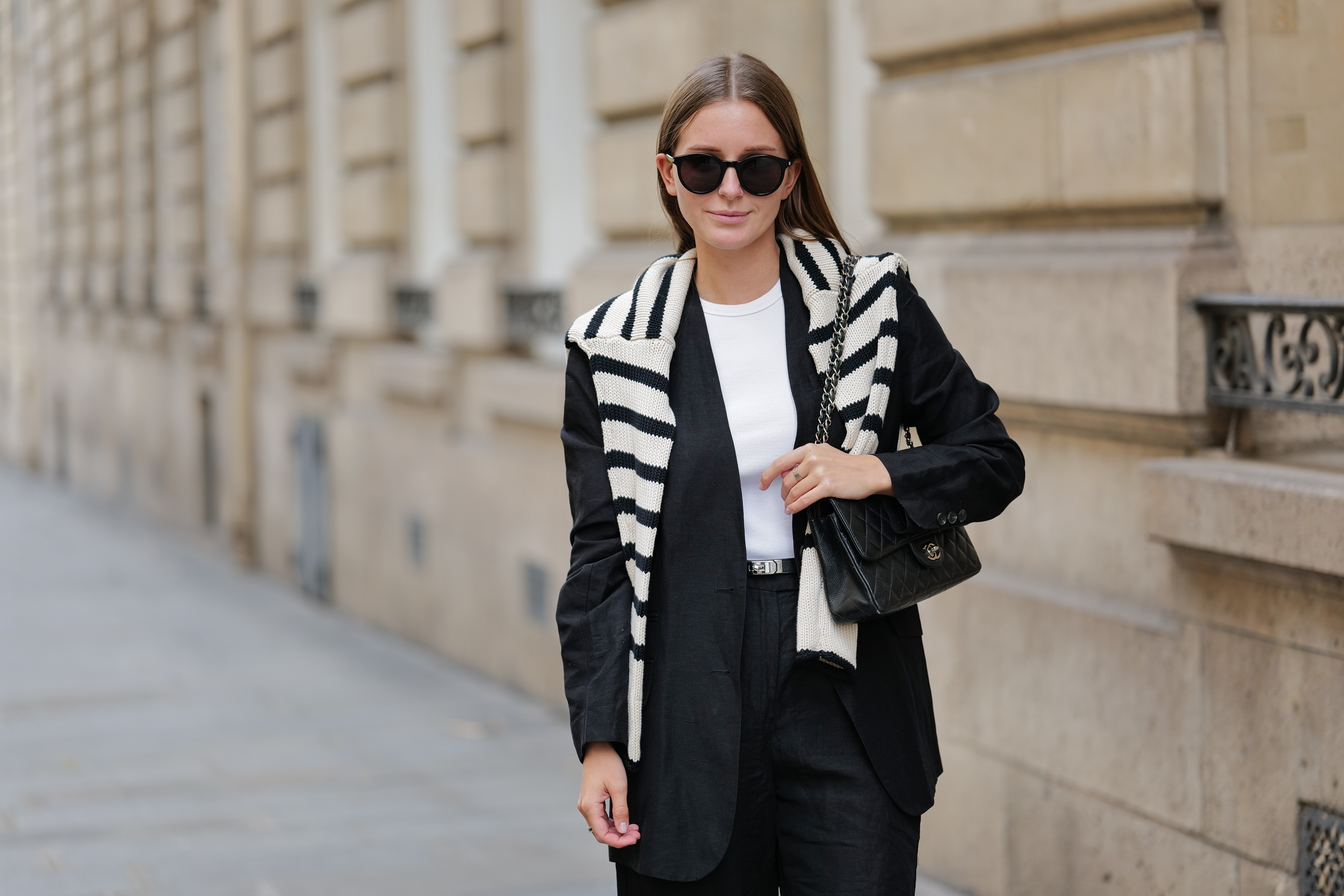 Diane Batoukina wears black large sunglasses, a white halter-neck tank-top from Anine Bing, a black blazer jacket from Massimo Dutti, a white latte and black striped print pattern wool pullover from Zara, a black shiny grained leather small Kelly belt from Hermes, black suit shorts from Massimo Dutti, a black shiny leather quilted Timeless handbag from Chanel, during a street style fashion photo session, on April 28, 2023 in Paris, France.