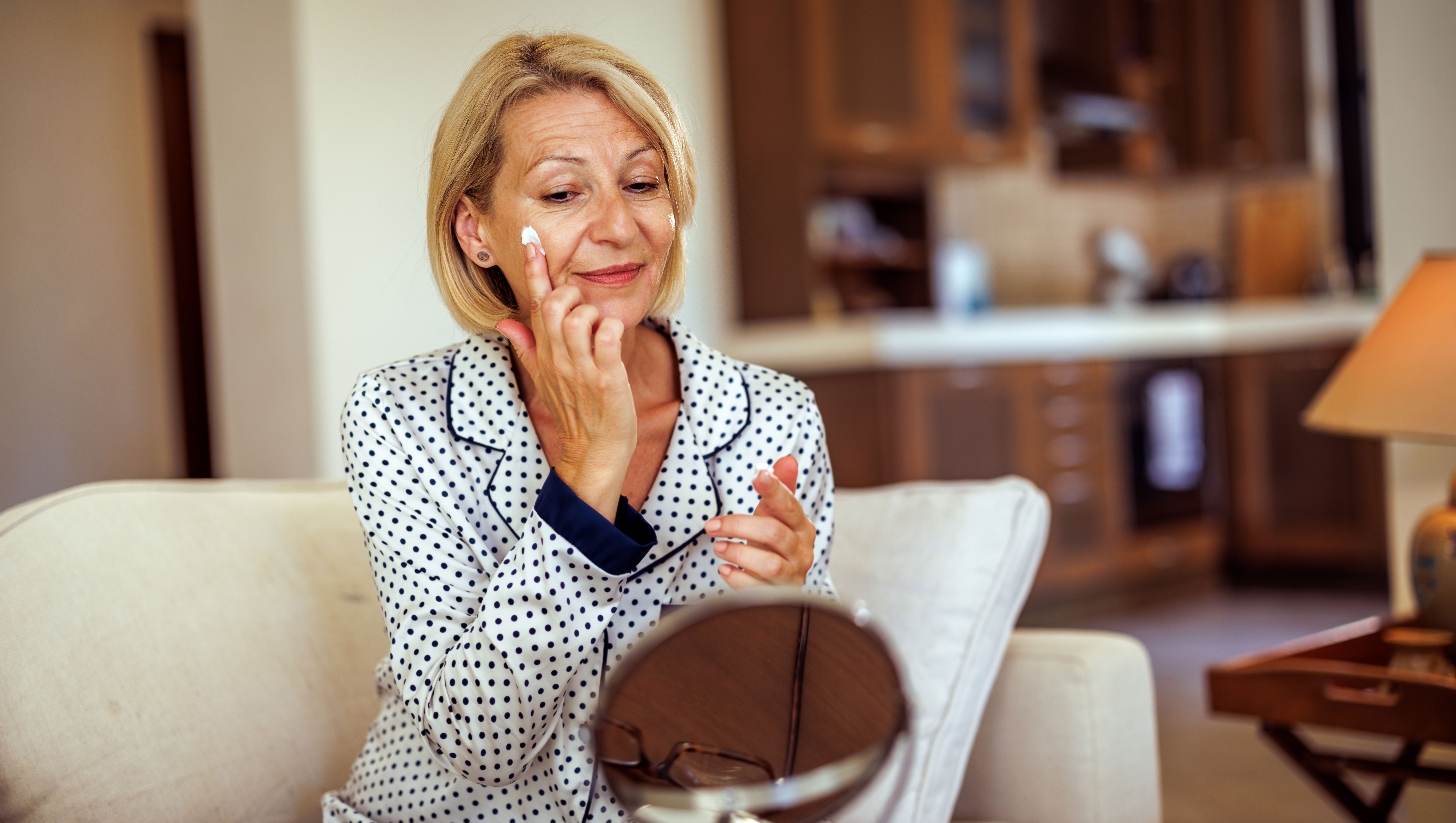 Smiling mature woman applying face cream under her eye while doing skin care routine at home - stock photo