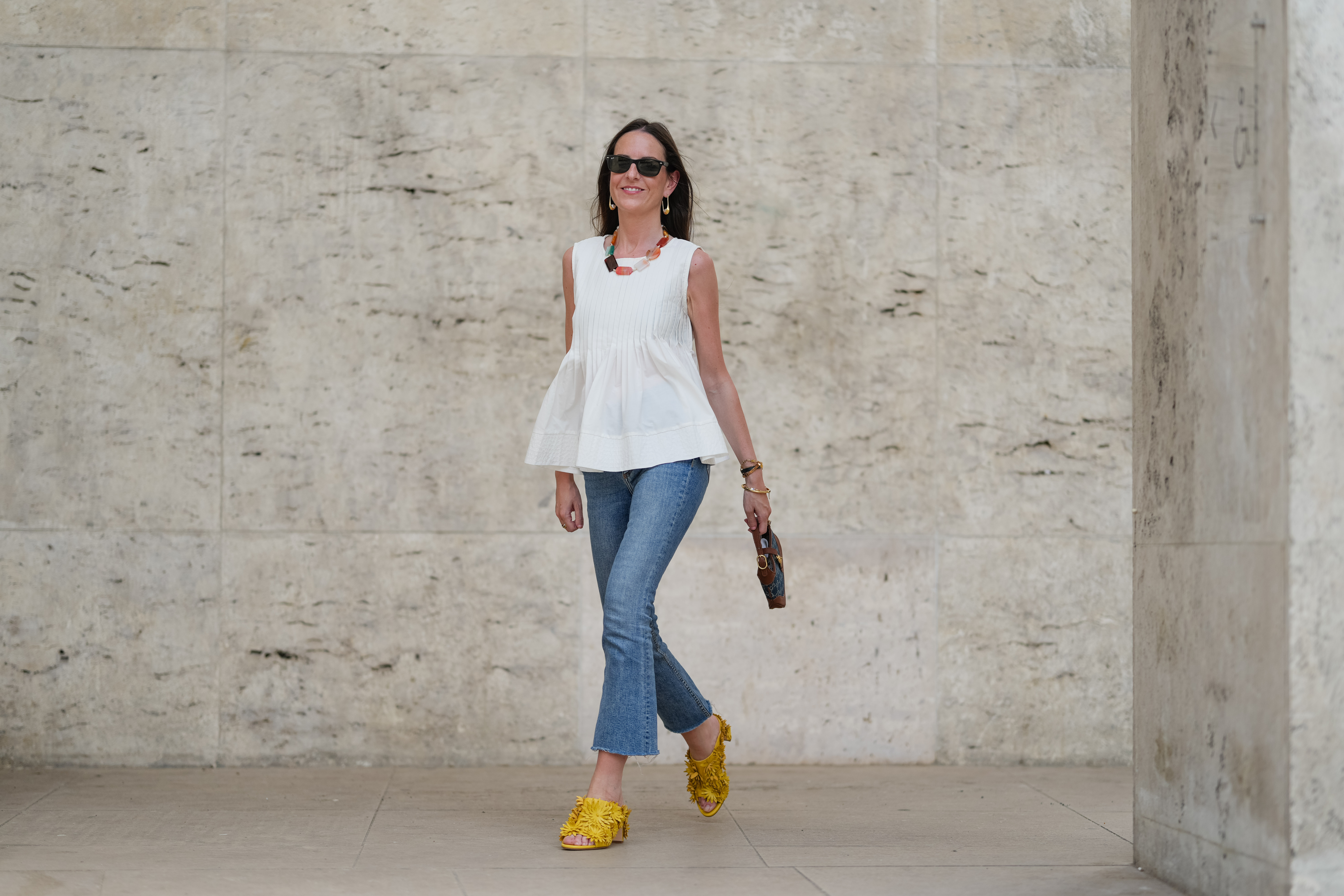 Alba Garavito Torre wears black sunglasses, long straight brown hair with a center part, a white pleated peplum sleeveless blouse by Commense, and blue high-rise raw-hem denim jeans. A yellow clog sandal with floral detailing by AGL is worn, paired with a jean denim and brown mini Jackie handbag by Gucci. Accessories include a chunky necklace with colorful natural stones by Sempiterno Paris and gold jewelry. Full body shot, during a street style fashion photo session, on May 02, 2025 in Paris, France.
