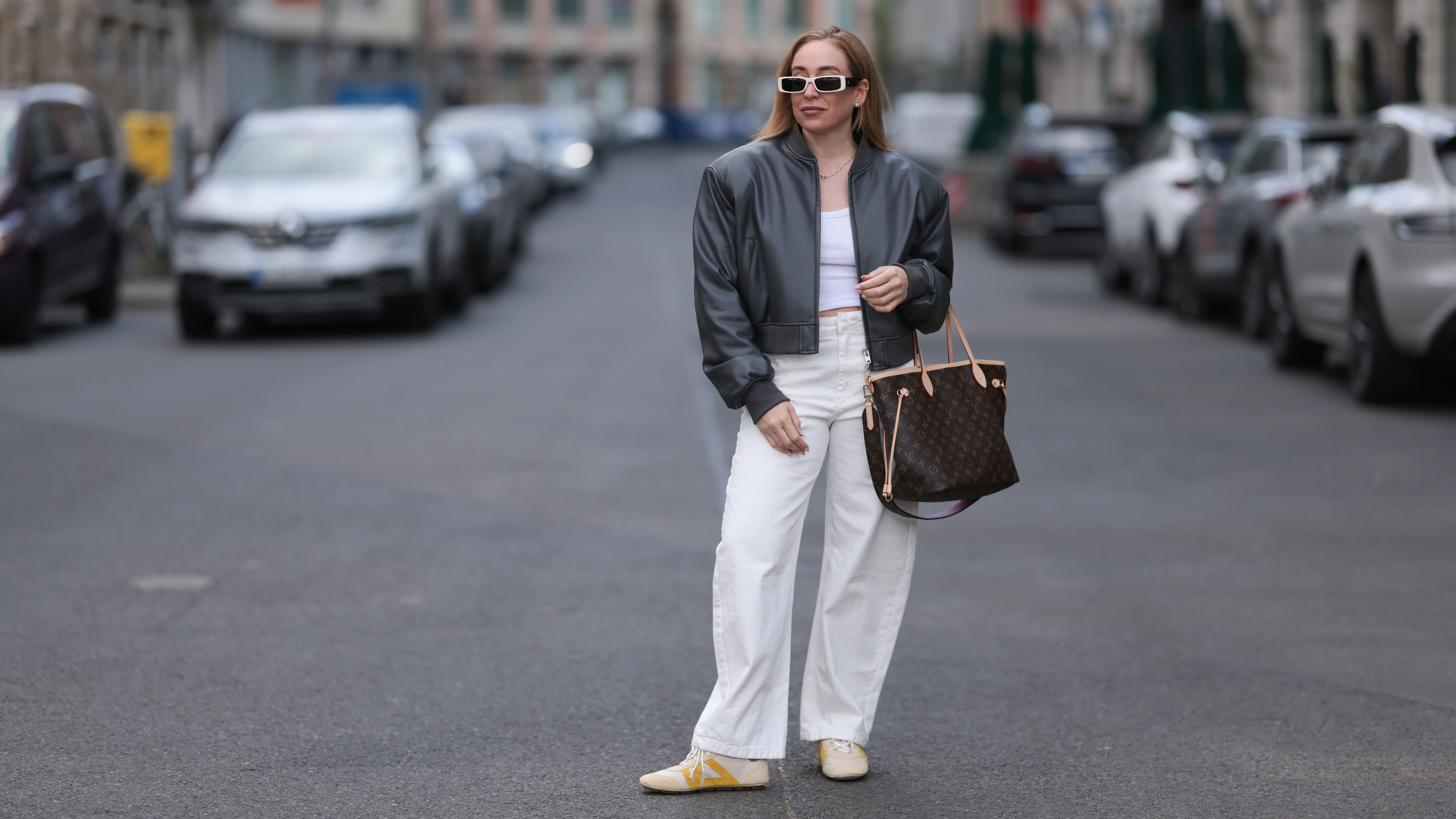 BERLIN, GERMANY - APRIL 09: Sonia Lyson seen wearing Linda Farrow bold white rectangular sunglasses that add a trendy touch, Zara white cotton fitted crop top, Zara high-waisted, wide-leg white relaxed jeans / denim pants, The Frankie Shop black, slightly glossy bomber jacket, Louis Vuitton brown Neverfull tote bag in the classic monogram print and Louis Vuitton beige Sneakerina sneakers / shoes with mustard yellow details, introducing a playful, sporty contrast to the otherwise minimalist outfit, on April 09, 2025 in Berlin, Germany. (Photo by Jeremy Moeller/Getty Images)