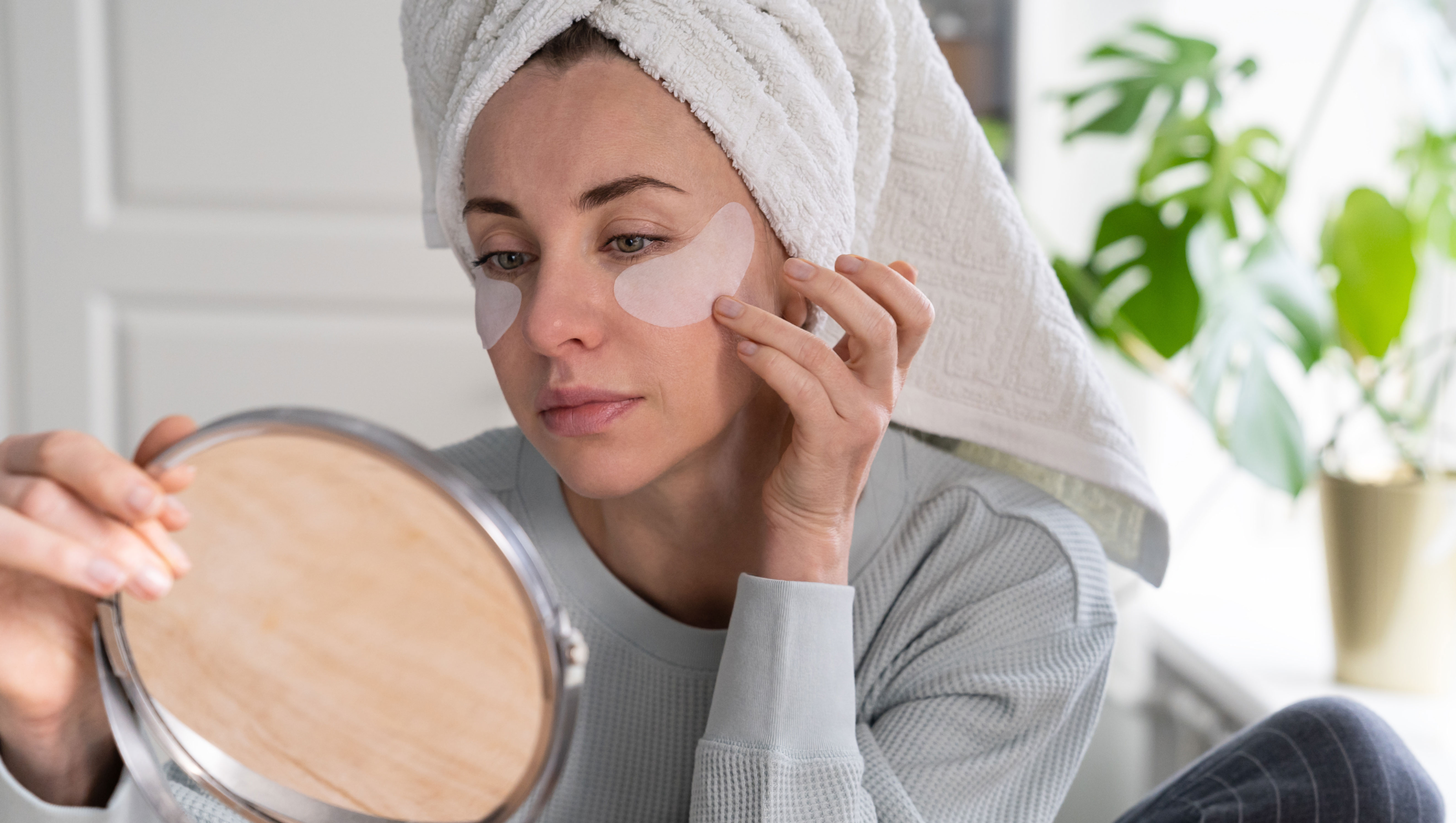 Woman with towel on head applying patches under eyes enriched with collagen, vitamin E, diminishes the signs of aging, helps reduse eye puffiness, looking in mirror. Face skin care beauty at home.