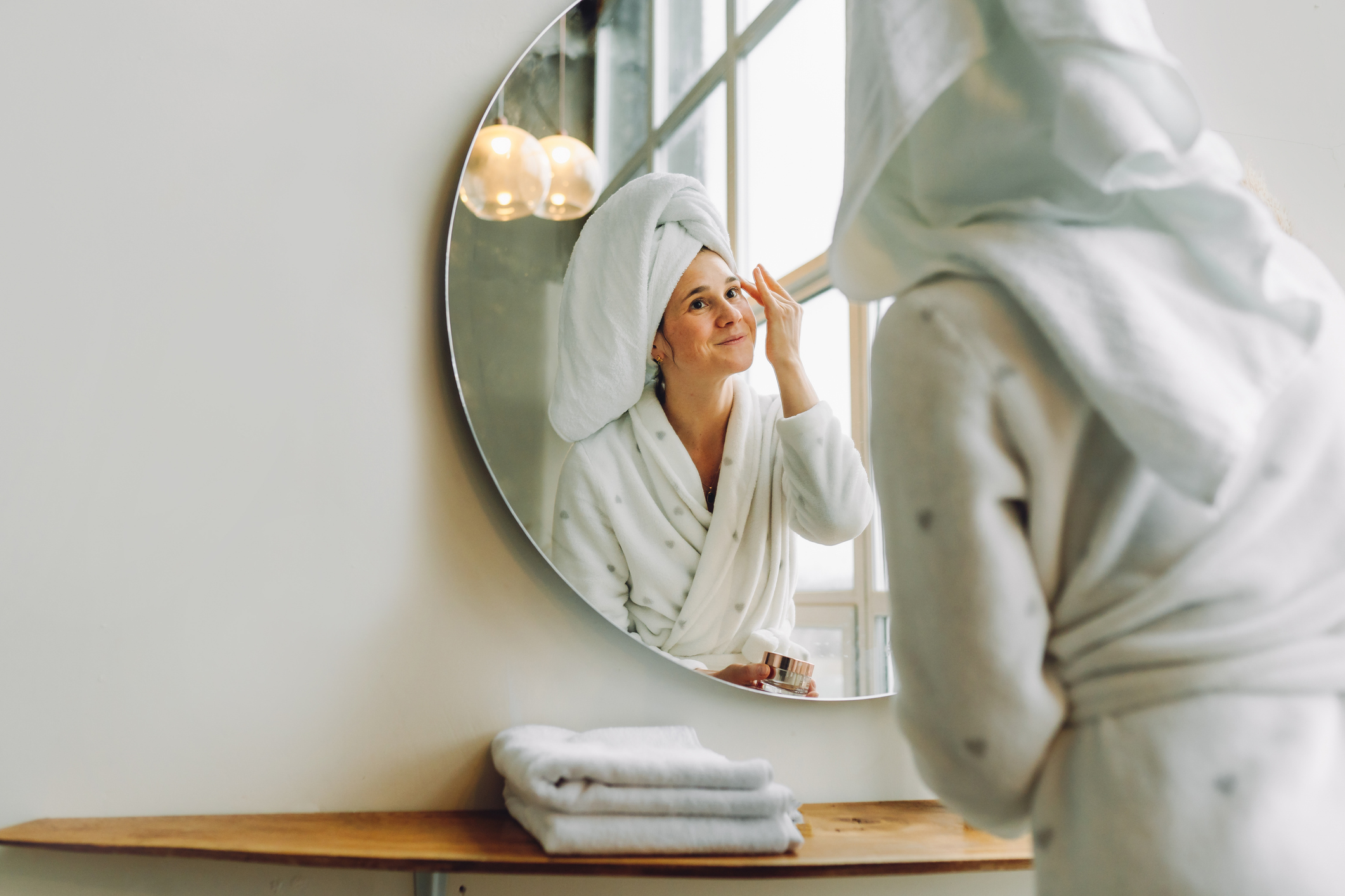Caucasian woman with towel on head applies moisturizer for hyperpigmentation in front of mirror in bright white bathroom.