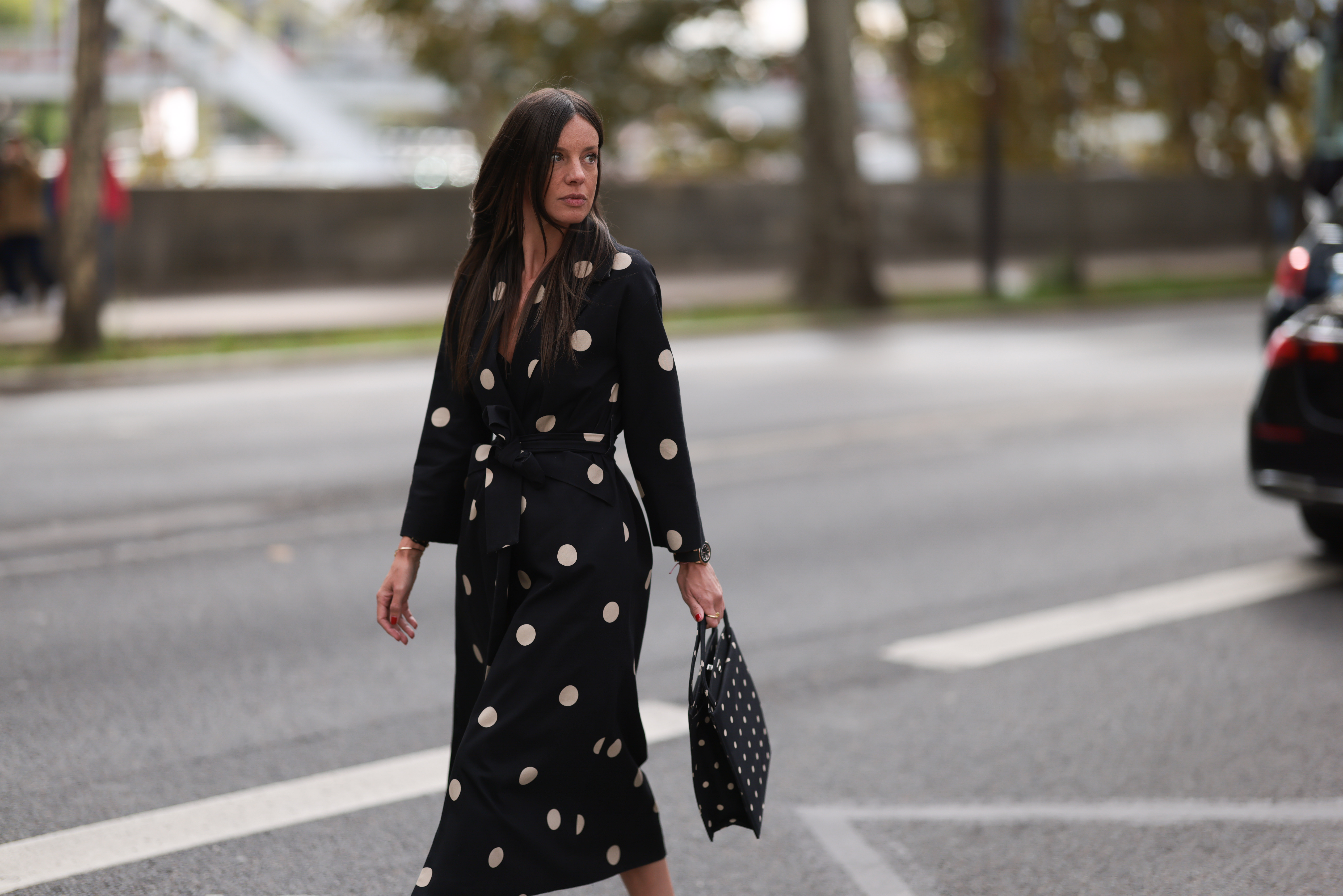 A Fashion Week guest is seen wearing round diamond stud earrings, a long-sleeved black midi dress with white polka dot pattern, notched lapels, and a tie belt; a gold bangle; a rose gold and black watch from Rolex; a gold ring; and a rectangular black handbag with a white polka dot pattern before the Elie Saab Womenswear Spring/Summer 2025 Show as part of Paris Fashion Week on September 28, 2024 in Paris, France.