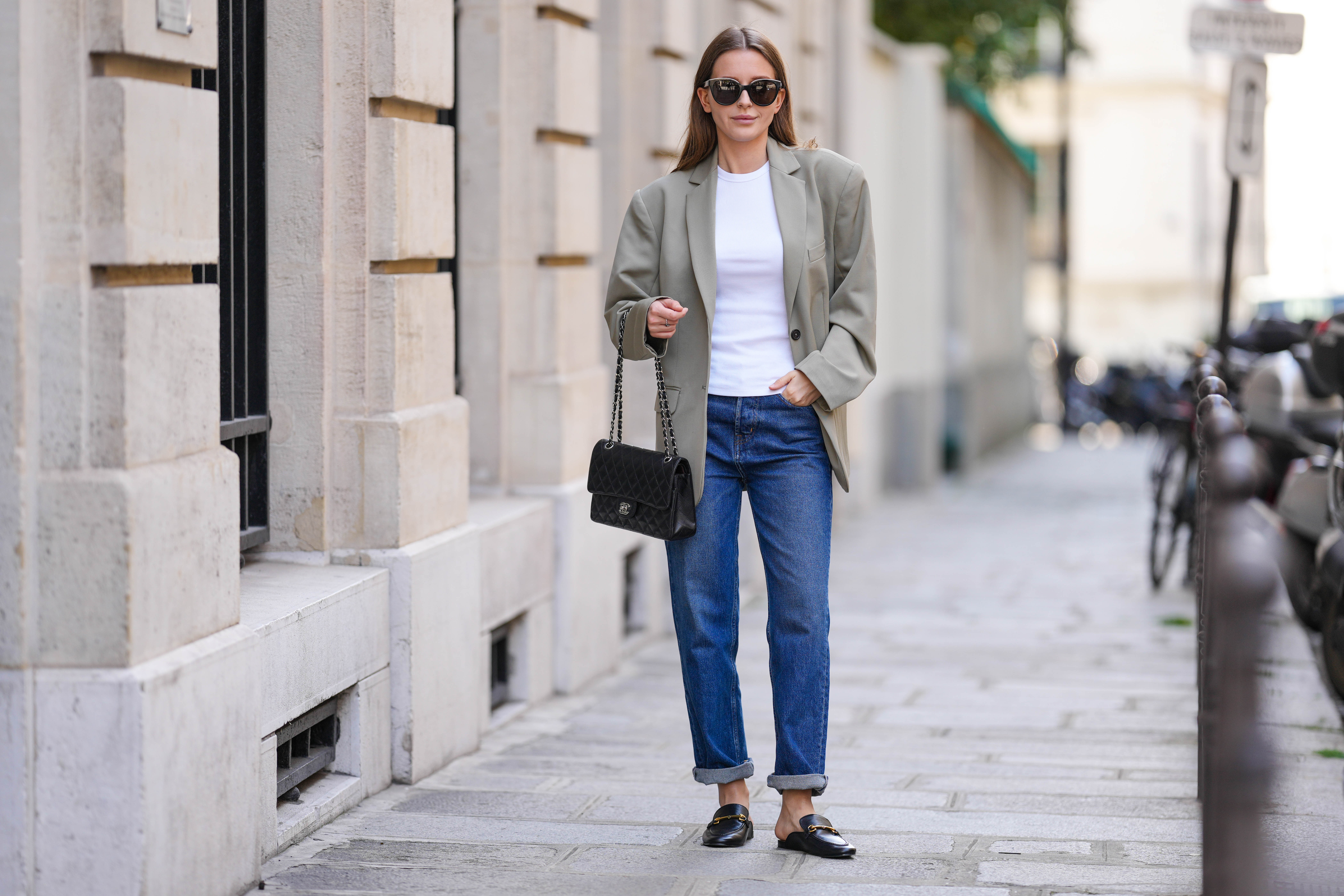 Diane Batoukina @diaanebt wears black sunglasses from Chanel, a white t-shirt, a beige gray oversized blazer jacket from Anine Bing, navy blue denim rolled-up large pants from Rouje, a black shiny grained leather Timeless handbag from Chanel, black shiny leather loafers mules from Massimo Dutti, during a street style fashion photo session, on April 15, 2022 in Paris, France.