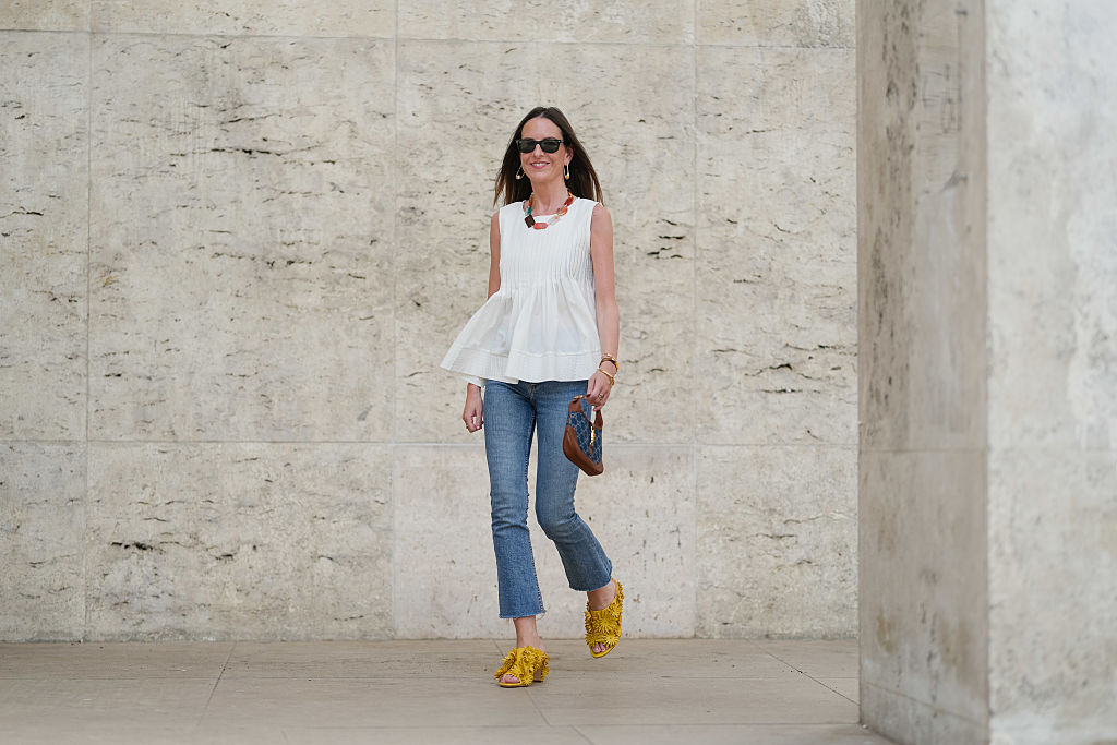 PARIS, FRANCE - MAY 02: Alba Garavito Torre wears black sunglasses, long straight brown hair with a center part, a white pleated peplum sleeveless blouse by Commense, and blue high-rise raw-hem denim jeans. A yellow clog sandal with floral detailing by AGL is worn, paired with a jean denim and brown mini Jackie handbag by Gucci. Accessories include a chunky necklace with colorful natural stones by Sempiterno Paris and gold jewelry. Full body shot, during a street style fashion photo session, on May 02, 2025 in Paris, France. (Photo by Edward Berthelot/Getty Images)
