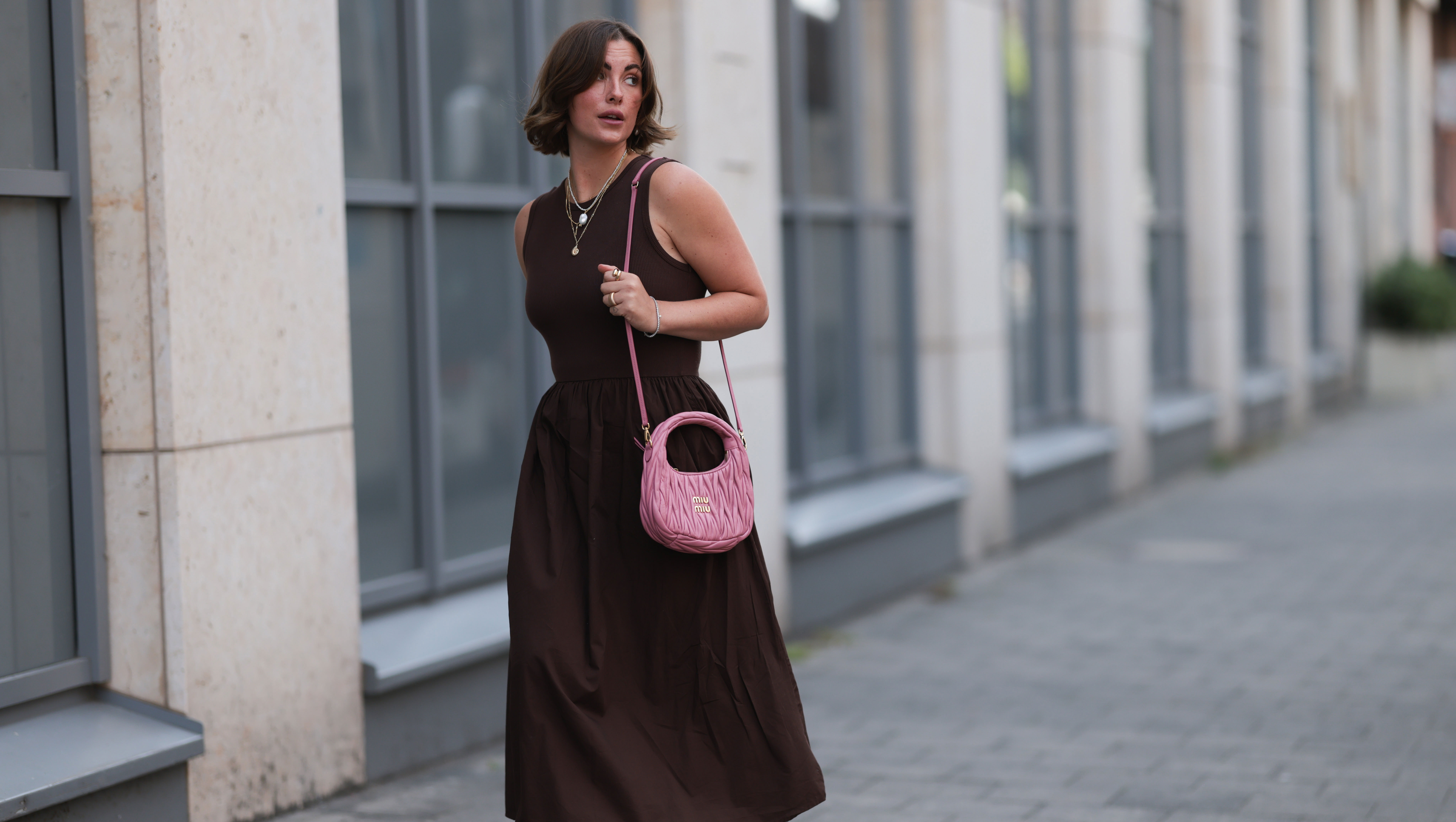 HAMBURG, GERMANY - JULY 18: Franzi Koenig is seen wearing several stud earrings, including gold seashell-shaped studs from fafe collection; layered gold necklaces with a seashell pendant, an F-letter pendant, and a white beaded necklace with a round organic-shaped pendant, also from fafe collection; a sleeveless dark brown midi dress with a ribbed top, high round neckline, and a gathered A-line skirt; a quilted pink leather “Wander” Hobo bag with gold logo hardware and a long thin shoulder strap from Miu Miu; several chunky gold rings, paired with delicate bracelets, both from fafe collection; and dark brown slip-on sandals with kitten heels on July 18, 2025 in Hamburg, Germany. (Photo by Jeremy Moeller/Getty Images)