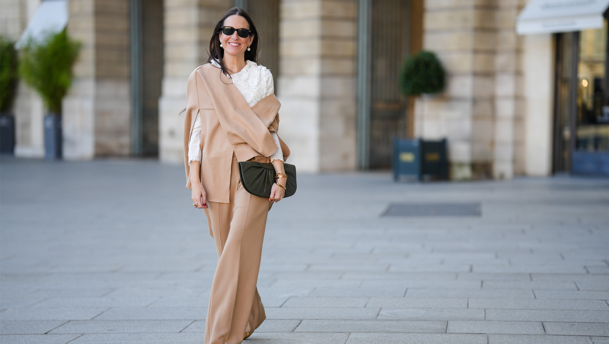 Alba Garavito Torre wears black sunglasses from Ray Ban, white large pearls earrings, a white shirt with cut-out lace pattern large collar, a beige felt / wool oversized cloak, matching beige wool / felt palazzo pants, a black shiny leather ruffled handbag, a gold Love bracelet from Cartier, gold bracelet, gold rings, brown leather pointed pumps heels shoes, during a street style fashion photo session, on October 28, 2021 in Paris, France