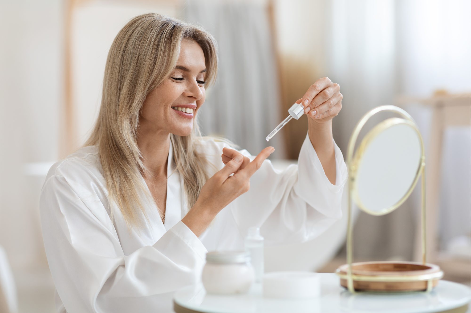 Happy beautiful middle aged woman in bathrobe sitting in front of mirror, holding bottle with facial oil or serum, lady applying face care product cosmetics after morning shower, copy space. (Photo by Prostock-Studio/Getty Images)