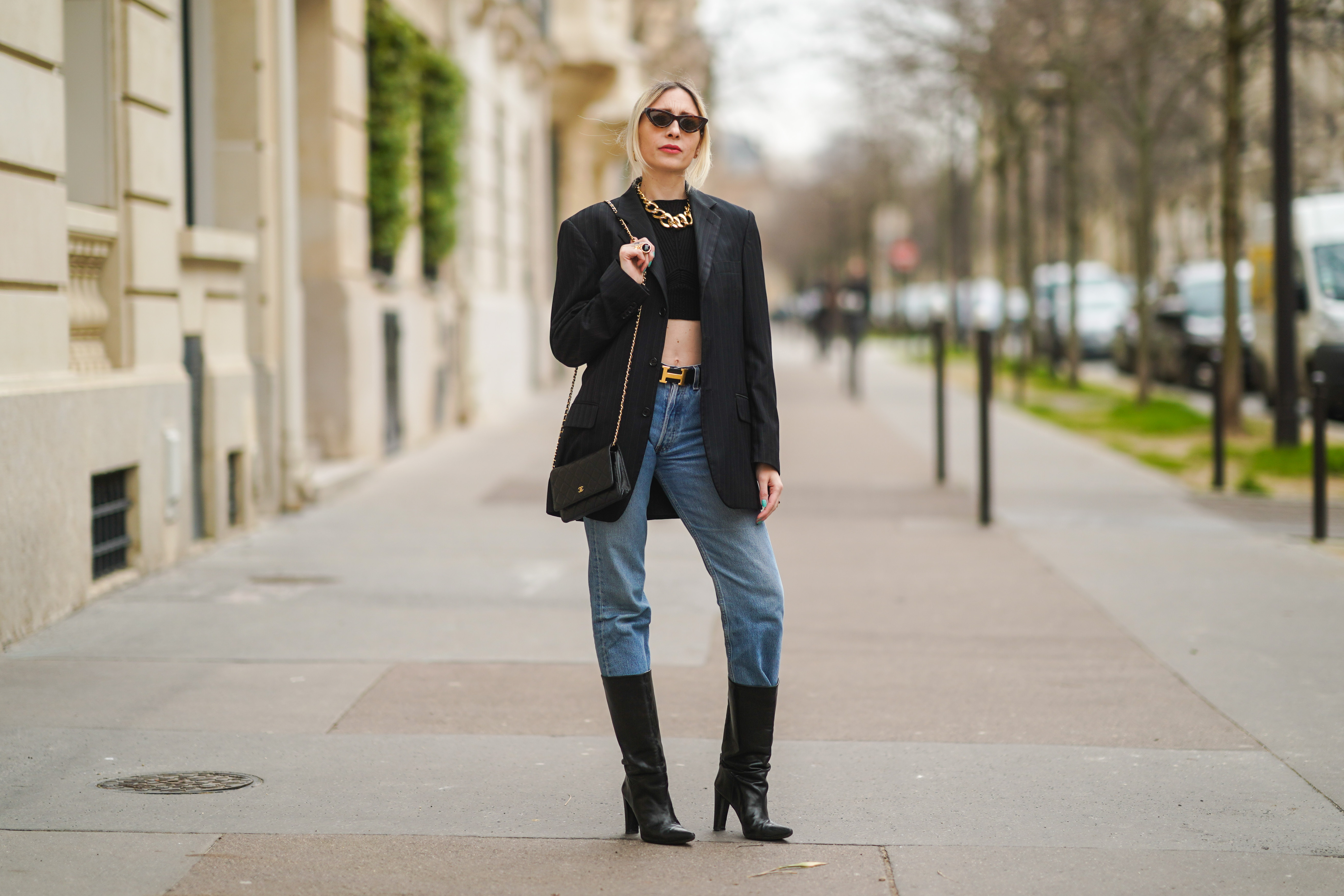 PARIS, FRANCE - FEBRUARY 22: Emy Venturini wears sunglasses, golden earrings, a golden large chain necklace, a black cropped top from Elisabetta Franchi, a black oversized blazer jacket, a Hermes belt, a black leather Chanel bag, blue denim jeans 501 Levi's, black high heeled pointy leather high boots, on February 22, 2021 in Paris, France.