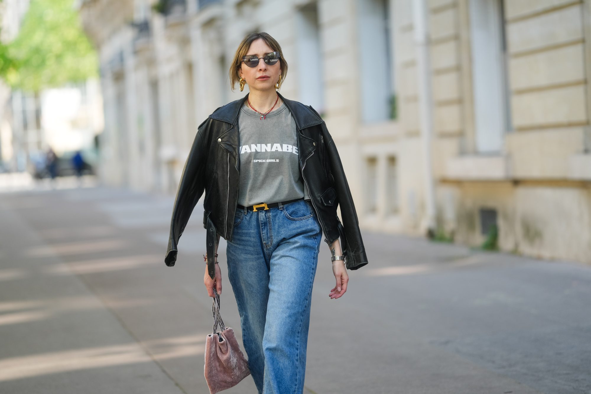 PARIS, FRANCE - APRIL 09: Emy Venturini wears a black leather jacket with wide lapels and zipped pockets, layered over a faded grey graphic T-shirt by Zara featuring “WANNABE” and the Spice Girls logo. Blue high-waisted jeans by The Frankie Shop and a black belt with a gold H buckle by Hermes are visible. Gold sculptural earrings by Rabanne and a red beaded necklace complete the look. Mid shot, during a street style fashion photo session, on April 09, 2025 in Paris, France. (Photo by Edward Berthelot/Getty Images)