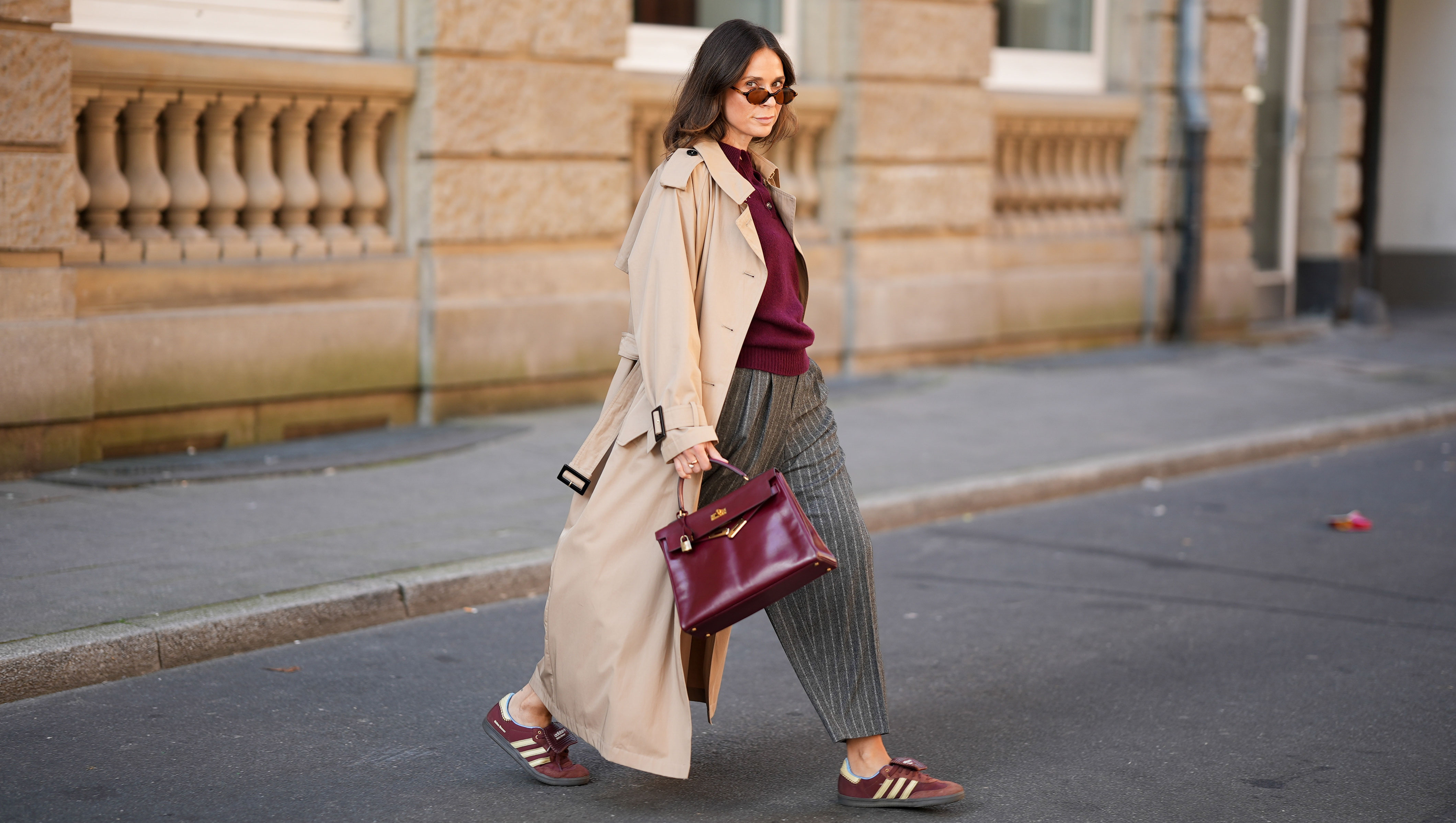 DUESSELDORF, GERMANY - AUGUST 19: Patricia Wirschke is seen wearing a Anna V Berg burgundy knit polo sweater with three buttons, collar and ribbed hem; a classic beige long Herno trench coat, with a notched collar, storm flap, and belted cuffs; a pair of high-waisted, wide-leg, ankle length Rossi pants that feature a gray pinstripe pattern with tailored pleats and loose silhouette; a pair of burgundy suede Adidas Samba X Wales Bonner sneakers with cream stripes; a structured, glossy burgundy leather Hermes Kelly bag with gold-tone hardware; a pair of Miu Miiu oval-shaped tortoiseshell sunglasses; her brown hair is styled in a straight, shoulder-length look with a middle part on August 19, 2025 in Duesseldorf, Germany. (Photo by Moritz Scholz/Getty Images)