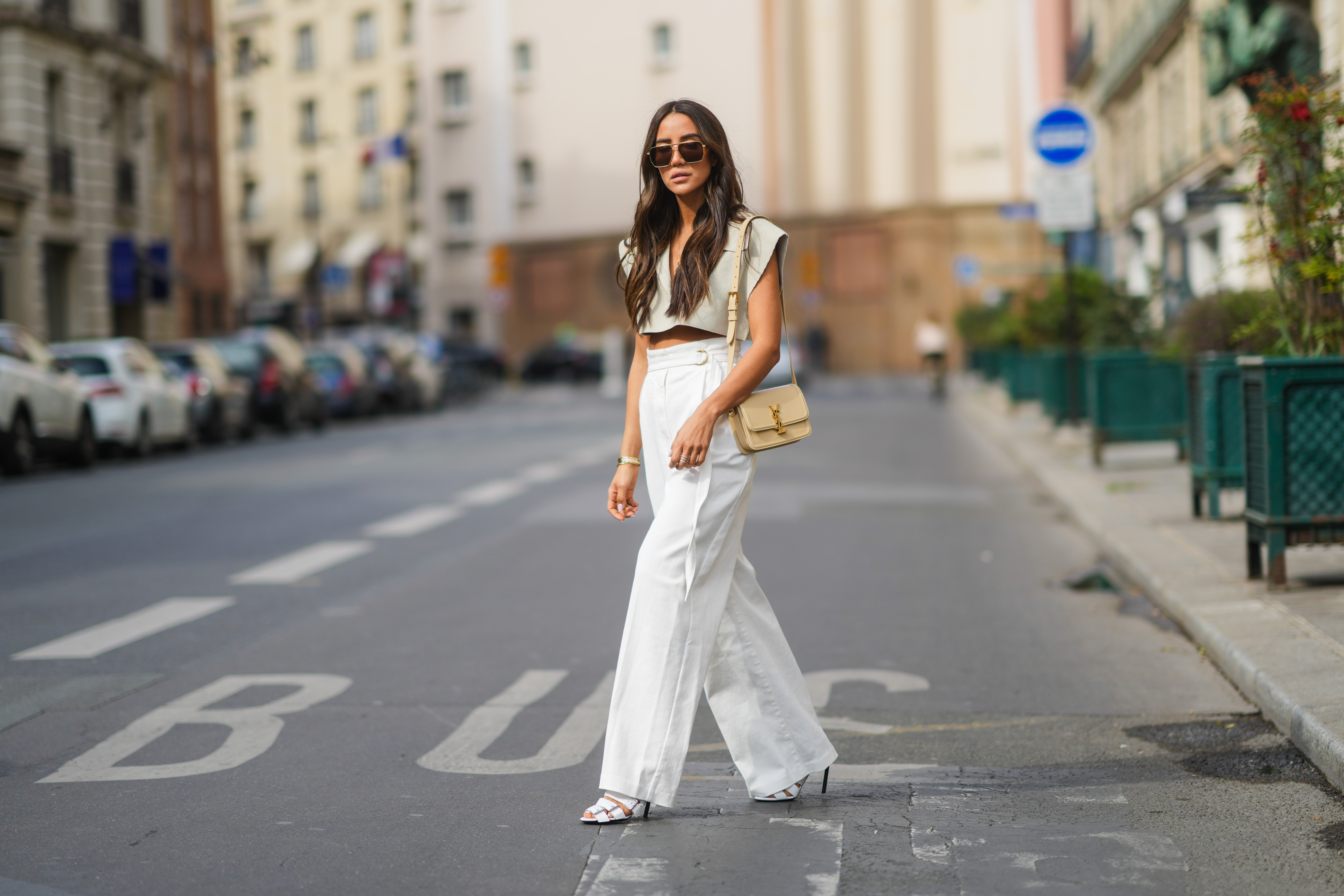 Tamara Kalinic wears gold Bottega Veneta sunglasses, a gold chain pendant necklace, a pale gray Jacquemus V-neck collar crop top with epaulets, white large flared Philosophy belted trousers pants, a gold Juste Un Clou Cartier bracelet, a gold Love Cartier bracelet, gold and silver Bee My Love Chaumet bracelets, a gold ring, a beige shiny leather Saint Laurent YSL shoulder bag, white shiny leather strappy heels Balenciaga sandals shoes, on April 29, 2021 in Paris, France.