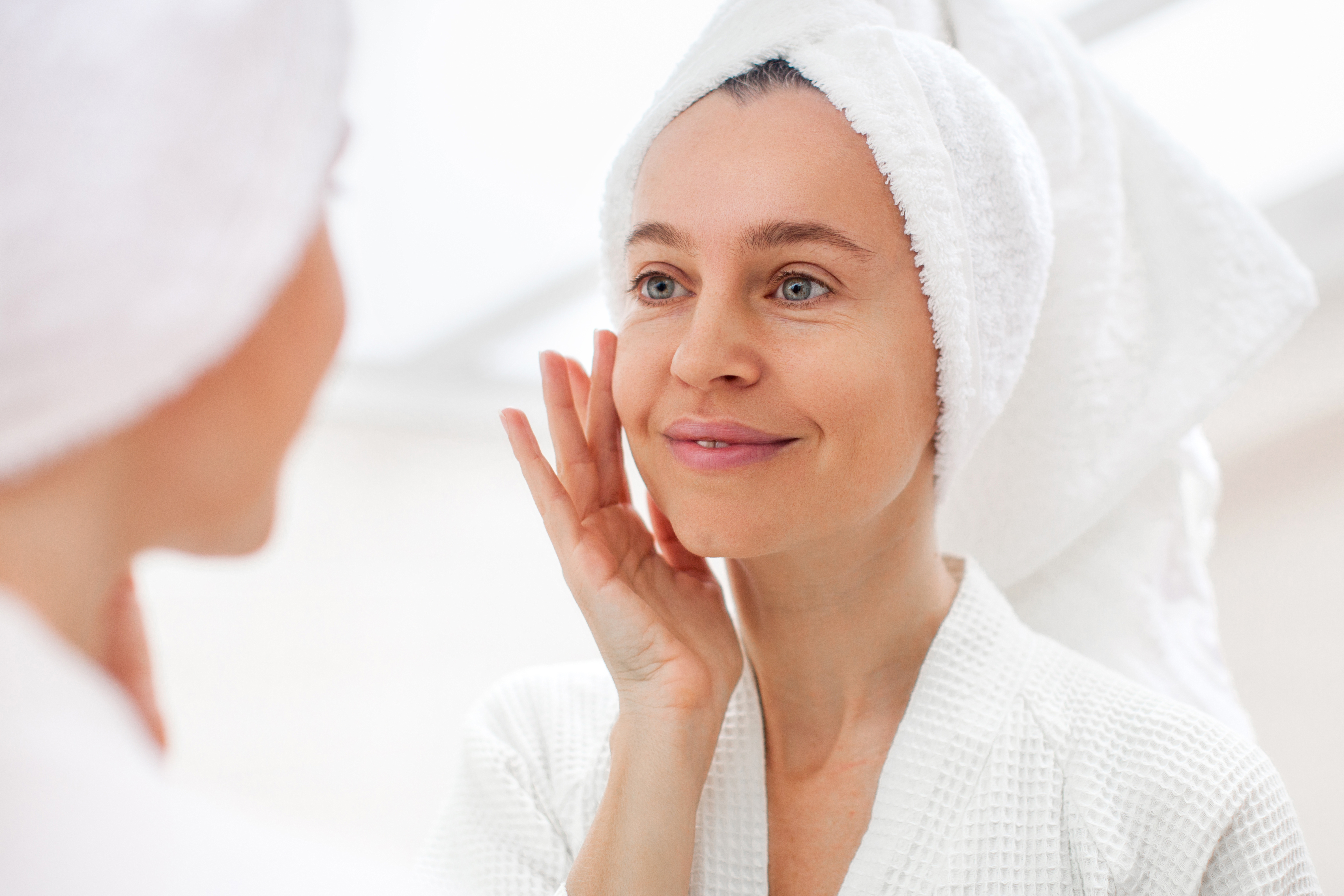 Happy 40s middle aged woman model touching face skin looking in mirror reflection. Smiling adult lady pampering, healthy moisturized skin care, aging beauty, skin care treatment cosmetics concept