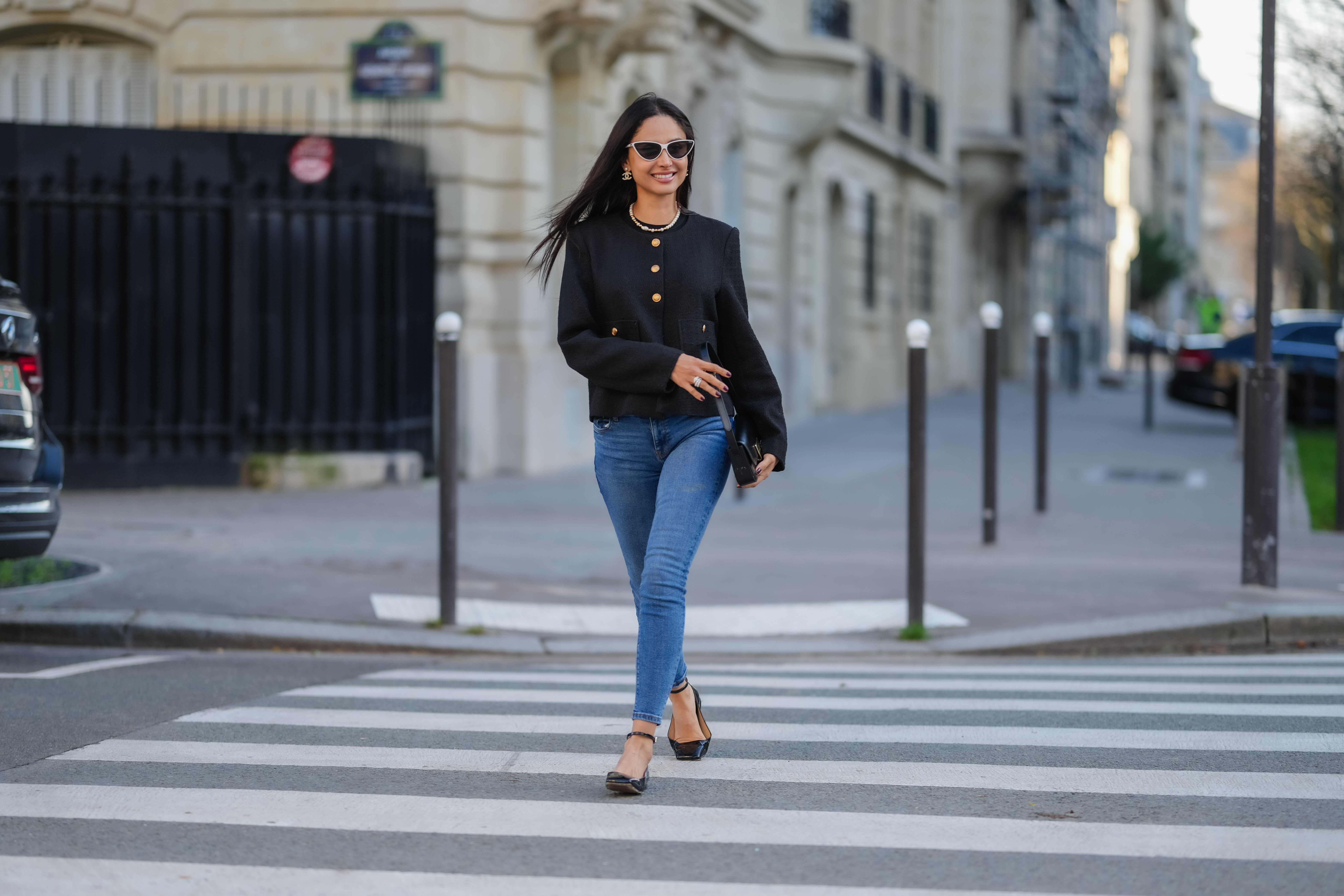 Patricia Gloria Conteras wears black and white sunglasses, silver Chanel earrings, a pearl necklace, a black jacket with gold buttons, blue denim jeans / pants, a black leather bag, black leather ballerinas shoes, during a street style fashion photo session, on January 07, 2025 in Paris, France.