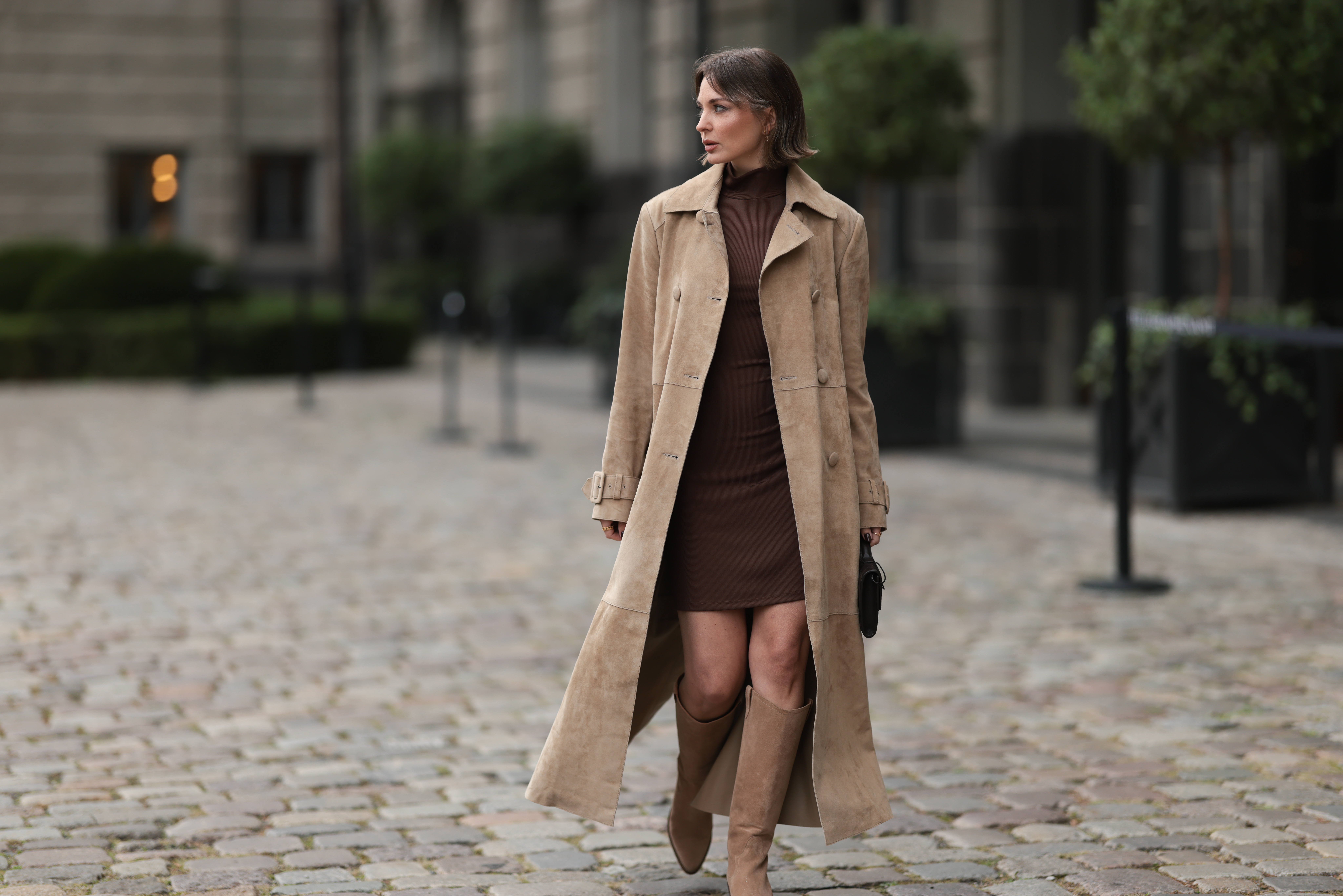 Yana Pries seen wearing a long suede leather trench coat from NOUR HAMMOUR in a warm beige tone. The coat features double-breasted buttons, a structured collar, and belted cuffs, creating a luxurious yet timeless silhouette. Her hair is cut into a sleek bob, light brown with subtle highlights, styled with a middle part and softly curved ends that frame her face. Underneath she wears Even&Odd fitted chocolate-brown mini dress, with a high neckline, Bottega Veneta Andiamo clutch, a structured black leather bag with a gold knot detail on the strap and Bobbies knee-high Bobbies suede leather boots in a soft beige tone, on September 17, 2025 in Berlin, Germany.