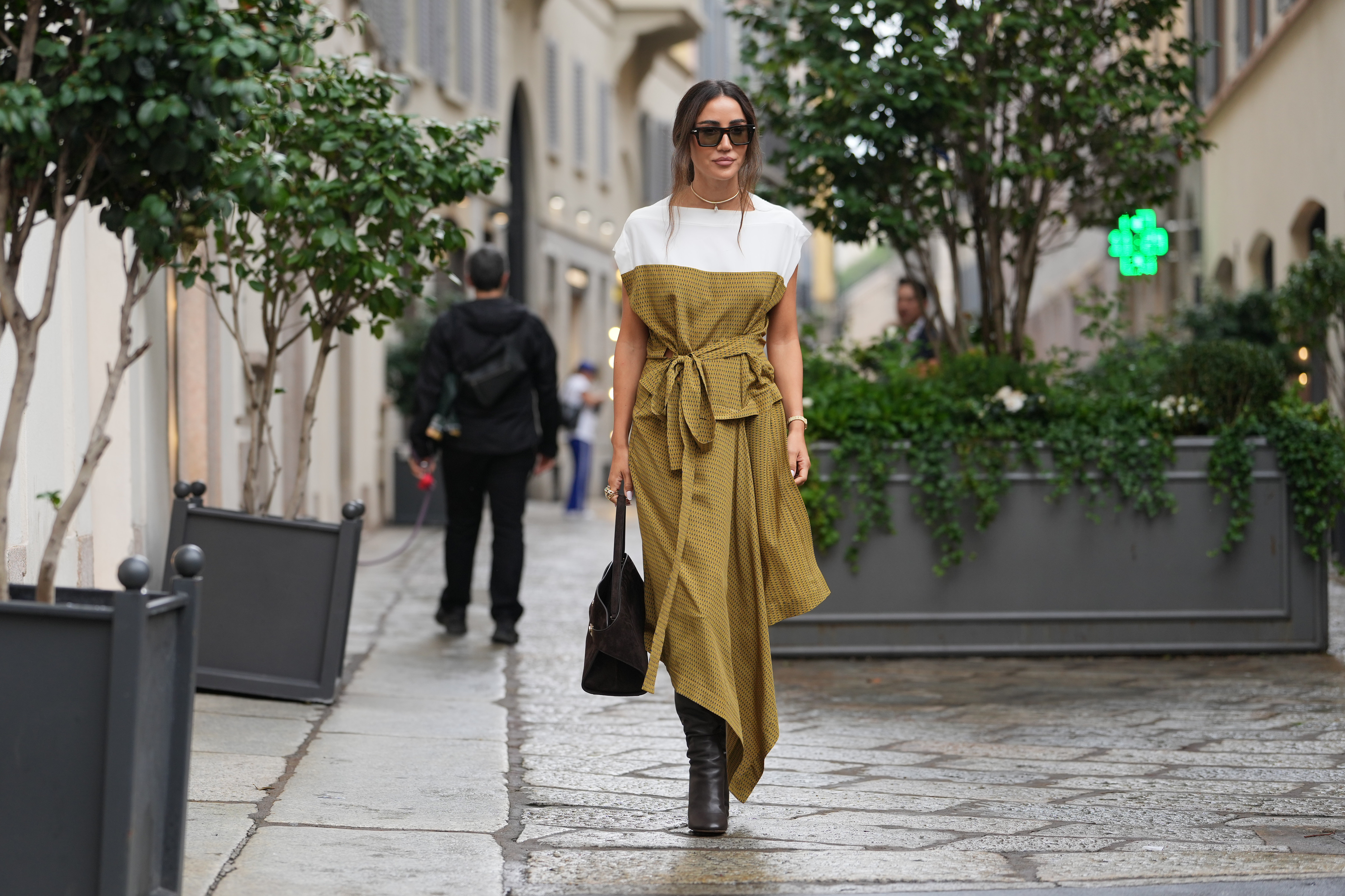 MILAN, ITALY - SEPTEMBER 27: Tamara Kalinic wears long dark brown hair styled in a low loose ponytail with a center part, black rectangular sunglasses, a gold choker-length necklace with matching gold bracelet and rings, a brown suede top-handle satchel bag, a mustard yellow patterned midi dress with a white shoulder yoke and capped sleeves, a self-tie waist belt and an asymmetrical handkerchief hem in a lightweight woven fabric, dark brown leather knee-high boots with a stacked high heel, outside Ferragamo, during Milan Fashion Week - Womenswear Spring/Summer 2026, on September 27, 2025 in Milan, Italy (Photo by Edward Berthelot/Getty Images)