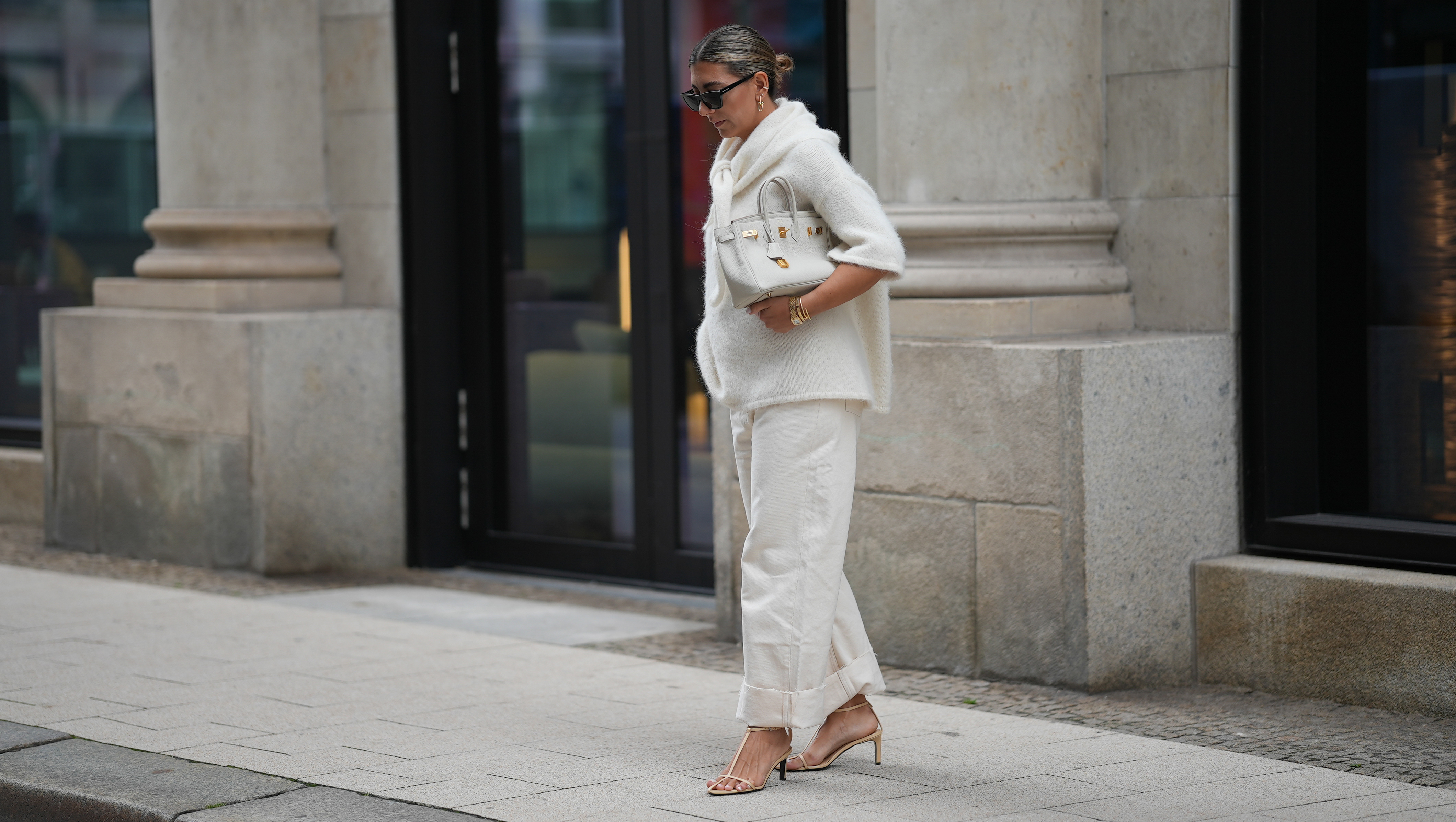 HAMBURG, GERMANY - SEPTEMBER 19: Aylin K√∂nig is seen wearing a short-sleeved, fluffy textured, oversized sweater in off-white with a high, tied neckline; relaxed off-white jeans with a wide cuff at the hem; nude strappy heeled sandals; a medium-sized white leather handbag with gold hardware; black rectangular sunglasses; gold link earrings from Tiffany & Co HardWear Collection; detailed gold bracelets from Cartier; a watch with a metallic wristband and white clock face from Cartier; her dark hair is pulled back into a neat, low bun on September 19, 2025 in Hamburg, Germany. (Photo by Moritz Scholz/Getty Images)