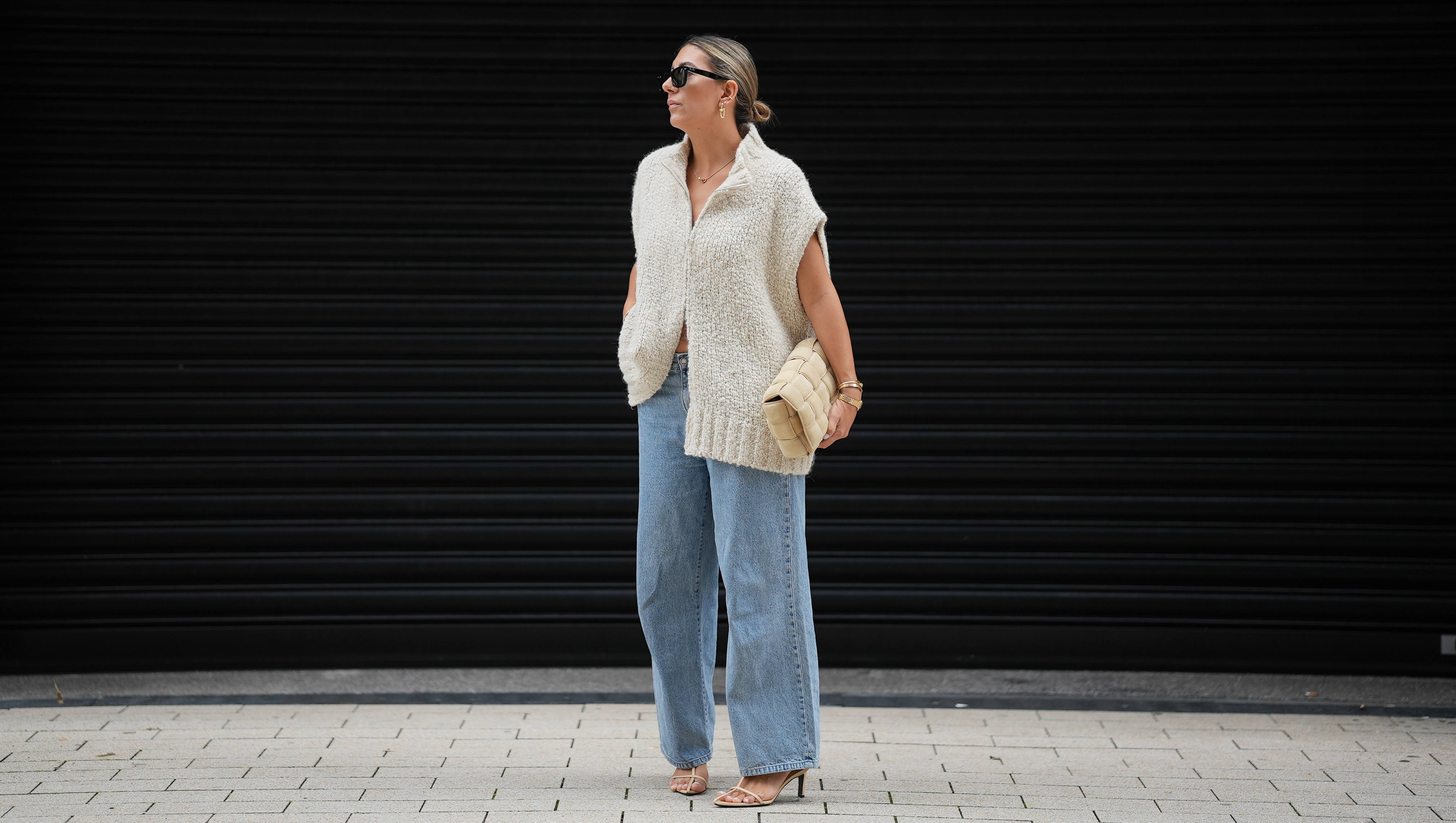 HAMBURG, GERMANY - SEPTEMBER 19: Aylin K&radic;&part;nig is seen wearing a loose, chunky knit sleeveless, zip-up sweater in a soft beige tone with a wide collar and relaxed drape; light blue straight-leg jeans with a slightly wide fit; nude strappy heeled sandals; a beige woven-style clutch bag with a padded texture; stacked gold bracelets from Cartier; a watch with a metallic wristband and white clock face from Cartier; gold detailed rings; gold link earrings from Tiffany & Co HardWear Collection; black rectangular sunglasses; her dark hair is pulled back into a neat, low bun on September 19, 2025 in Hamburg, Germany. (Photo by Moritz Scholz/Getty Images)
